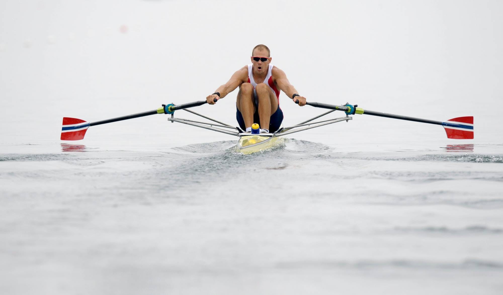 Roer i singelsculler med norske farger på årene ror på vannet. (Bildebeskrivelsen er laget av en KI-tjeneste)