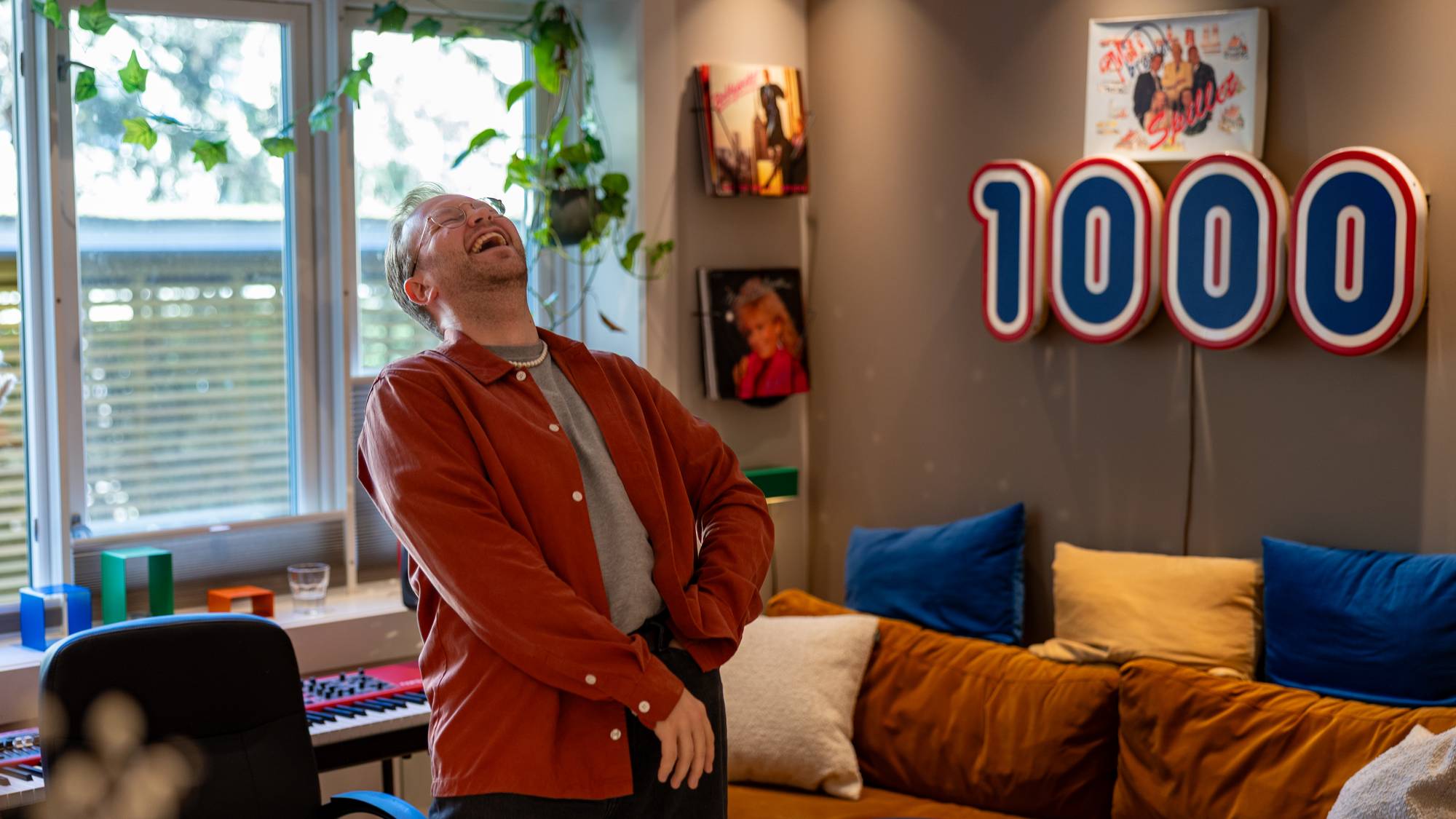 Mann med oransje skjorte og briller ler i ei leilegheit med sofa og eit stort Rema 1000-skilt i bakgrunnen