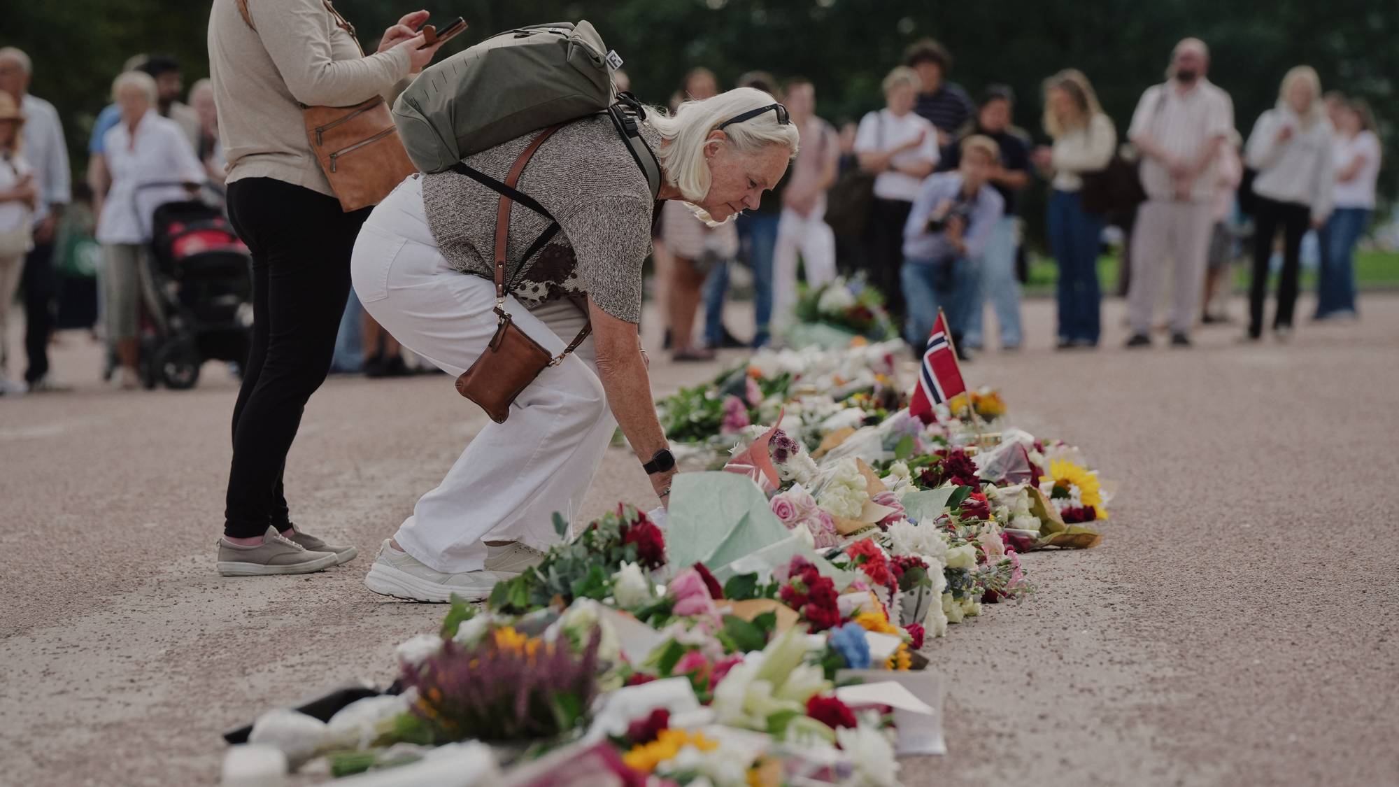 Kvinne legger ned blomster ved et minnested med mange blomster og et norsk flagg. Folk i bakgrunnen. 