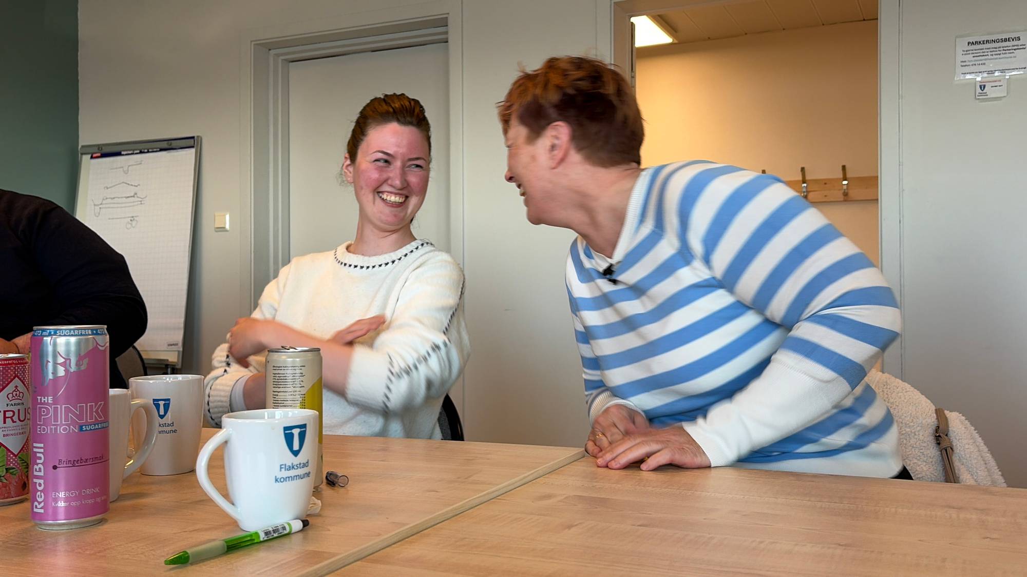 To kvinner sitter ved et bord. Den ene kvinnen til venstre ler og ser mot høyre. Hun har på seg en hvit genser med svarte detaljer. Den andre kvinnen til høyre ler og ser mot venstre. Hun har på seg en genser med blå og hvite striper. På bordet står det kopper med logoen til Flakstad kommune, en energidrikkboks og en penn. (Bildebeskrivelsen er laget av en KI-tjeneste)