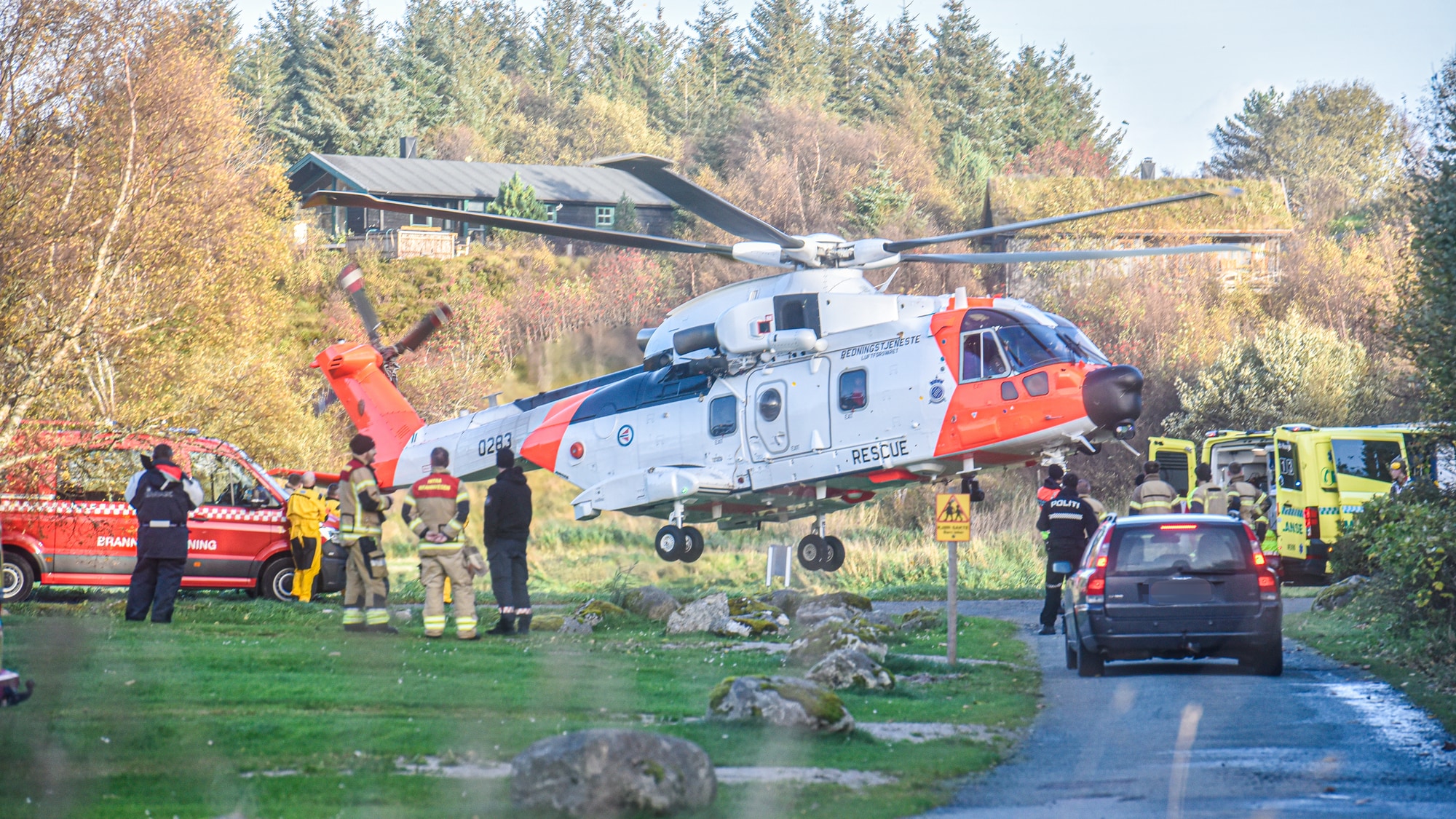 Et redningshelikopter letter. På bakken ser man ambulanse og en brannbil.