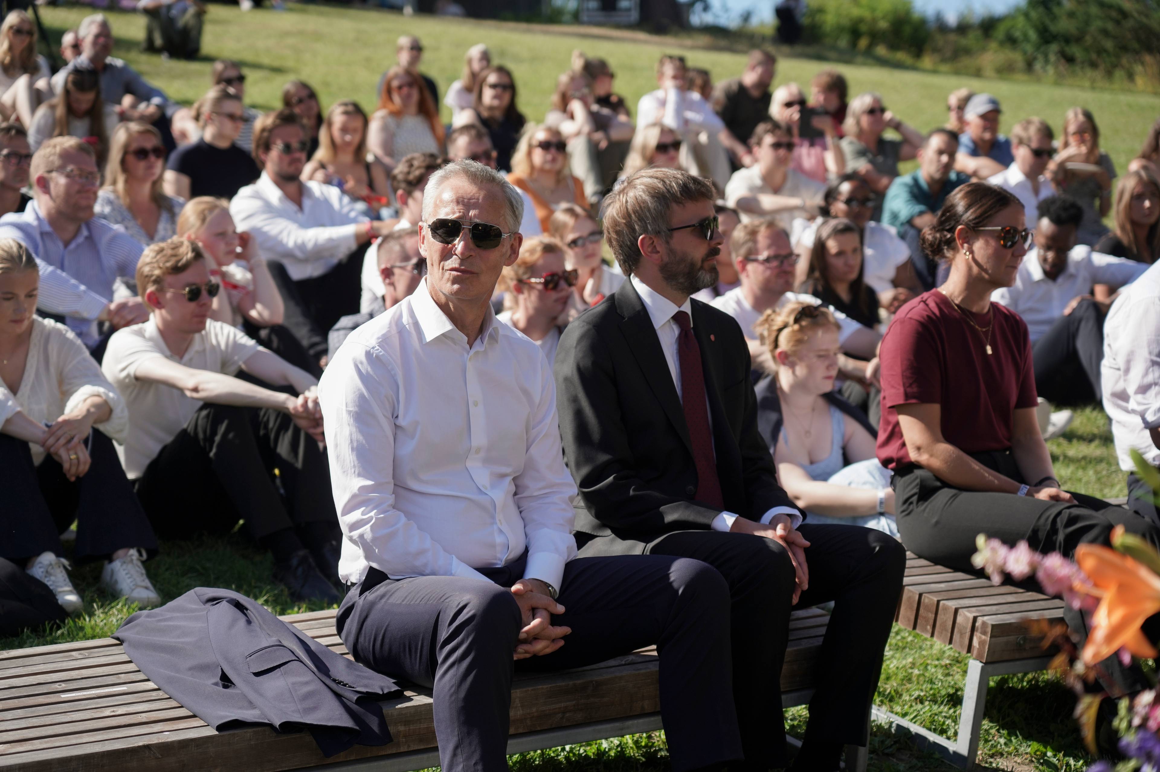 Jens Stoltenberg og andre sitter på benker foran et stort publikum i en utendørs forsamling. (Bildebeskrivelsen er laget av en KI-tjeneste)