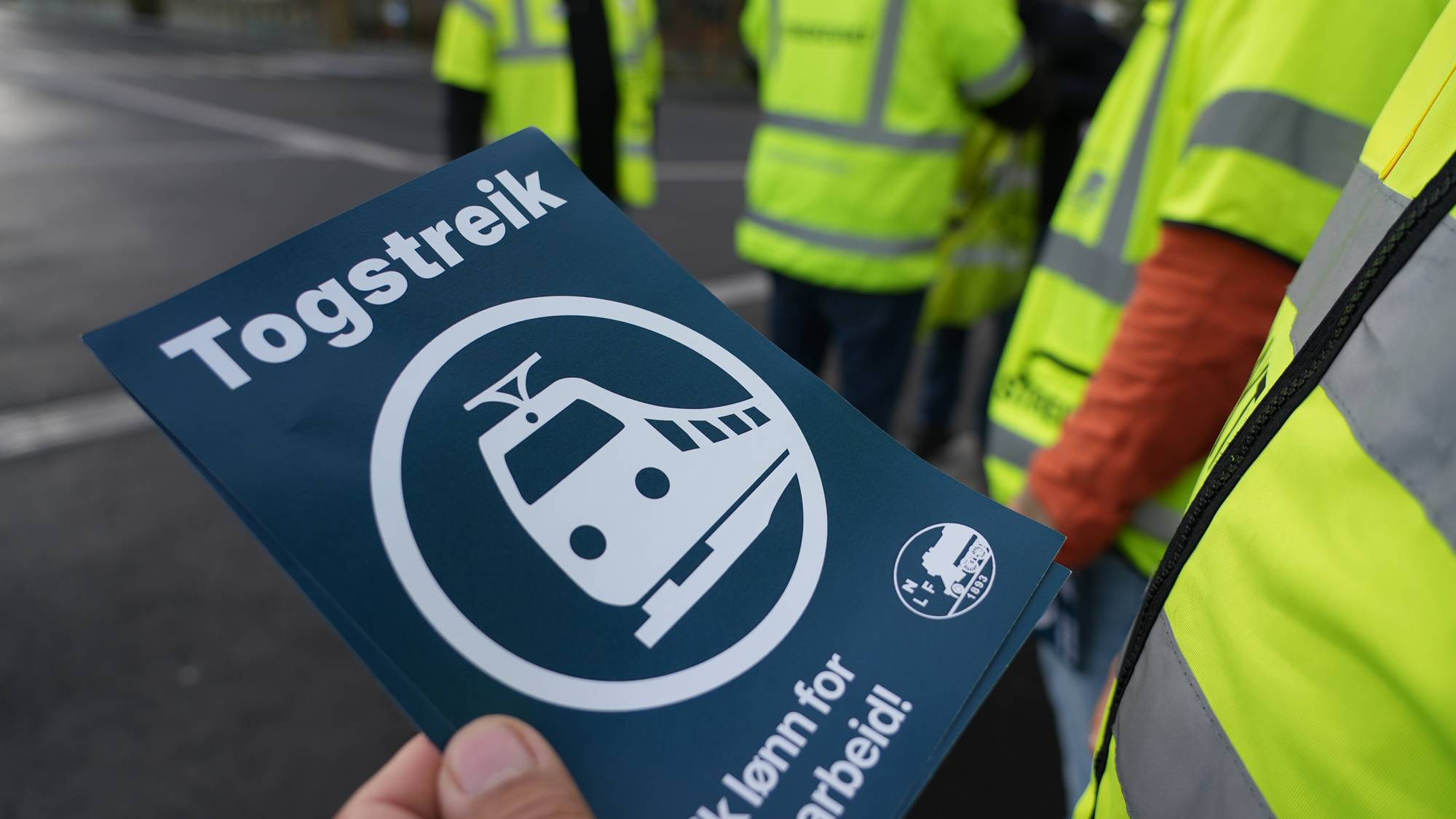 Person holder plakat med påskriften Togstreik foran personer i refleksvester. (Bildebeskrivelsen er laget av en KI-tjeneste)
