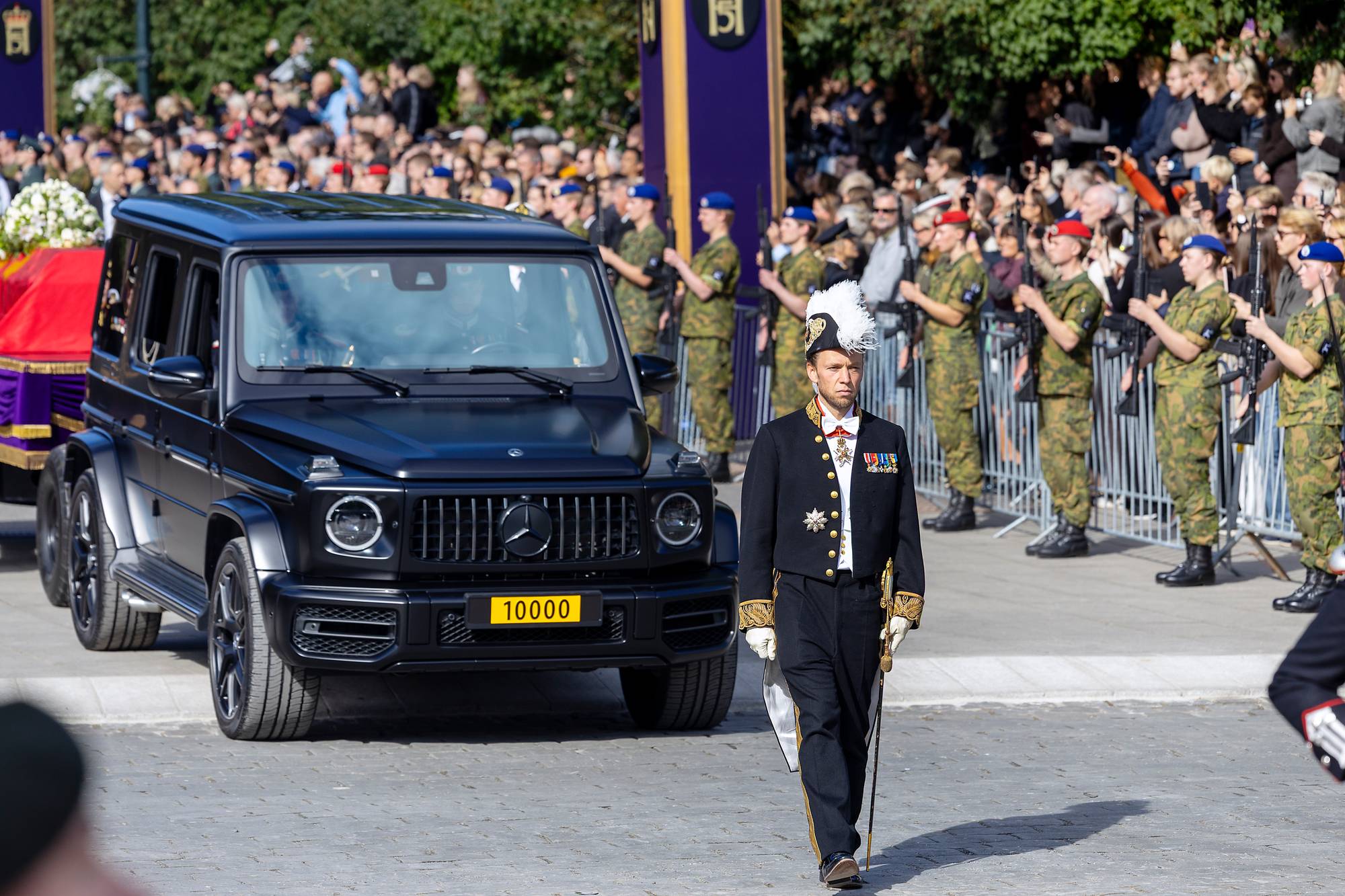 En offiser i uniform går foran en svart bil med et rødt kistelokk bak. Soldater står på vakt. (Bildebeskrivelsen er laget av en KI-tjeneste)