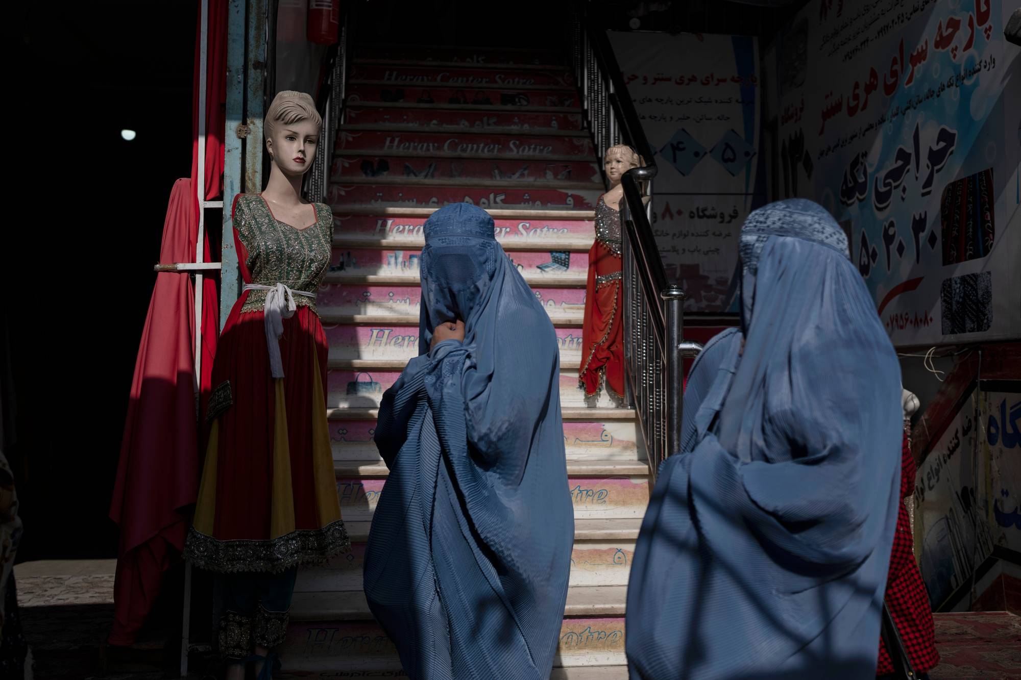 To kvinner i blå burka går opp en trapp utenfor en butikk. En mannequin med en rød og gul kjole står til venstre for trappen. Tekst på farsi er synlig på skiltene langs trappen og på veggen. (Bildebeskrivelsen er laget av en KI-tjeneste)