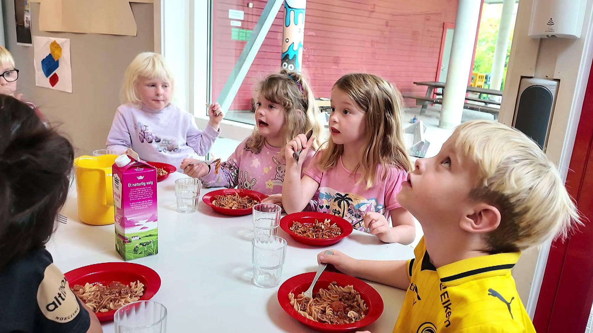 Barna sitter og spiser lunsj inne i barnehagen, alle guttene på bildet har på seg fotballdrakt, jentene er ikledd rosa. 