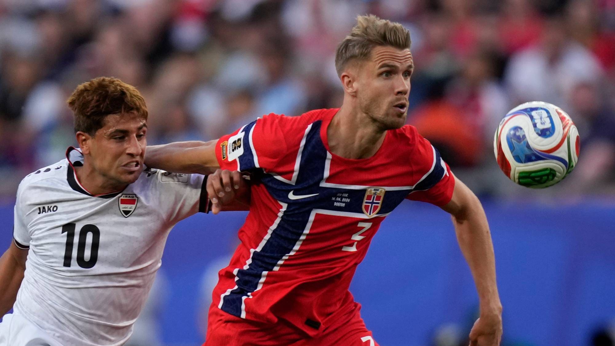 En norsk fotballspiller i rød drakt tar ballen fra en irakisk spiller i hvit drakt under en fotballkamp. Fotballspilleren med ballen er Martin Ødegaard. Kampen spilles på en bane med publikum i bakgrunnen. (Bildebeskrivelsen er laget av en KI-tjeneste)