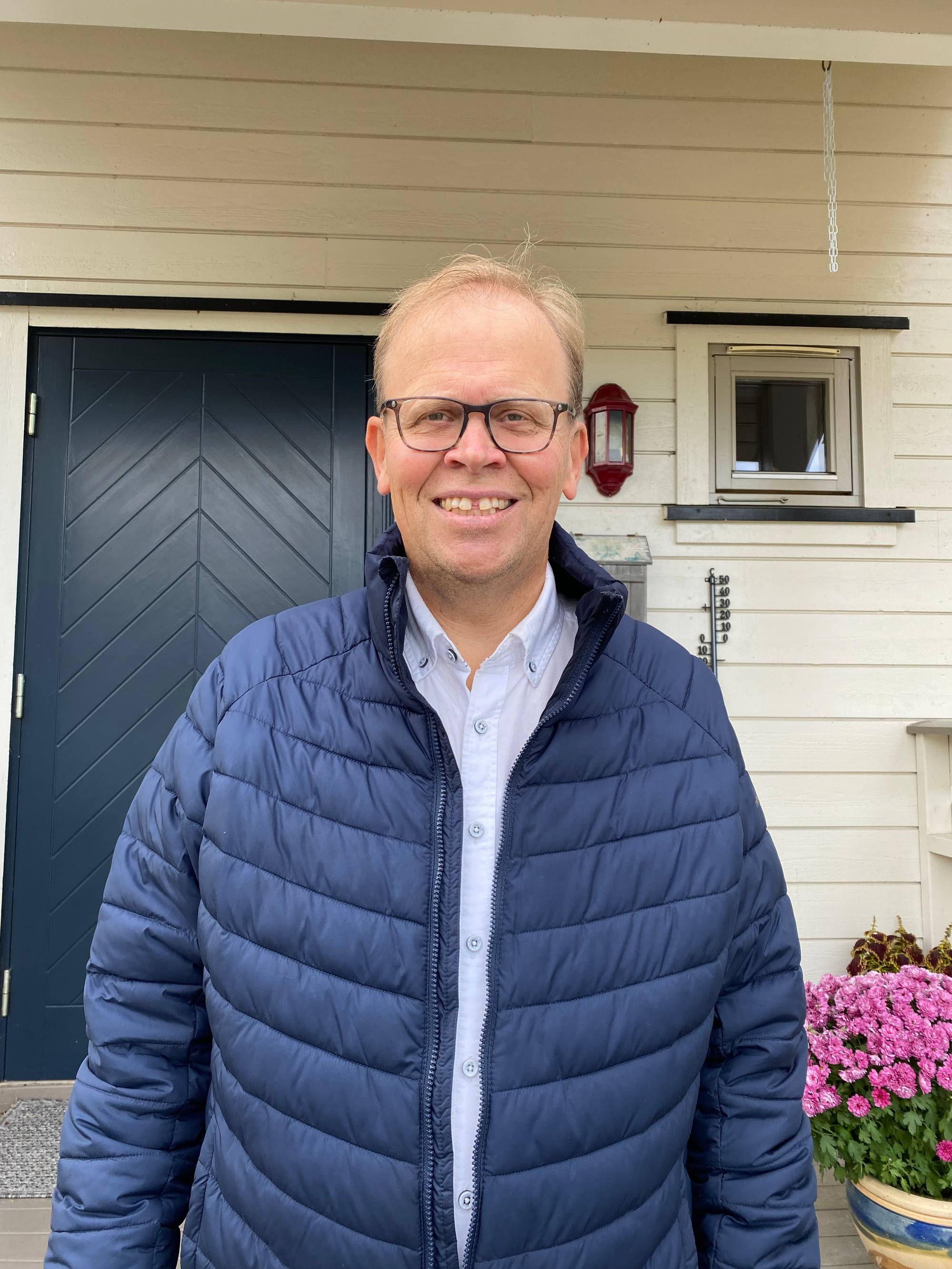 En mann med briller og blå boblejakke står foran en hvit husvegg og en mørk inngangsdør. (Bildebeskrivelsen er laget av en KI-tjeneste)