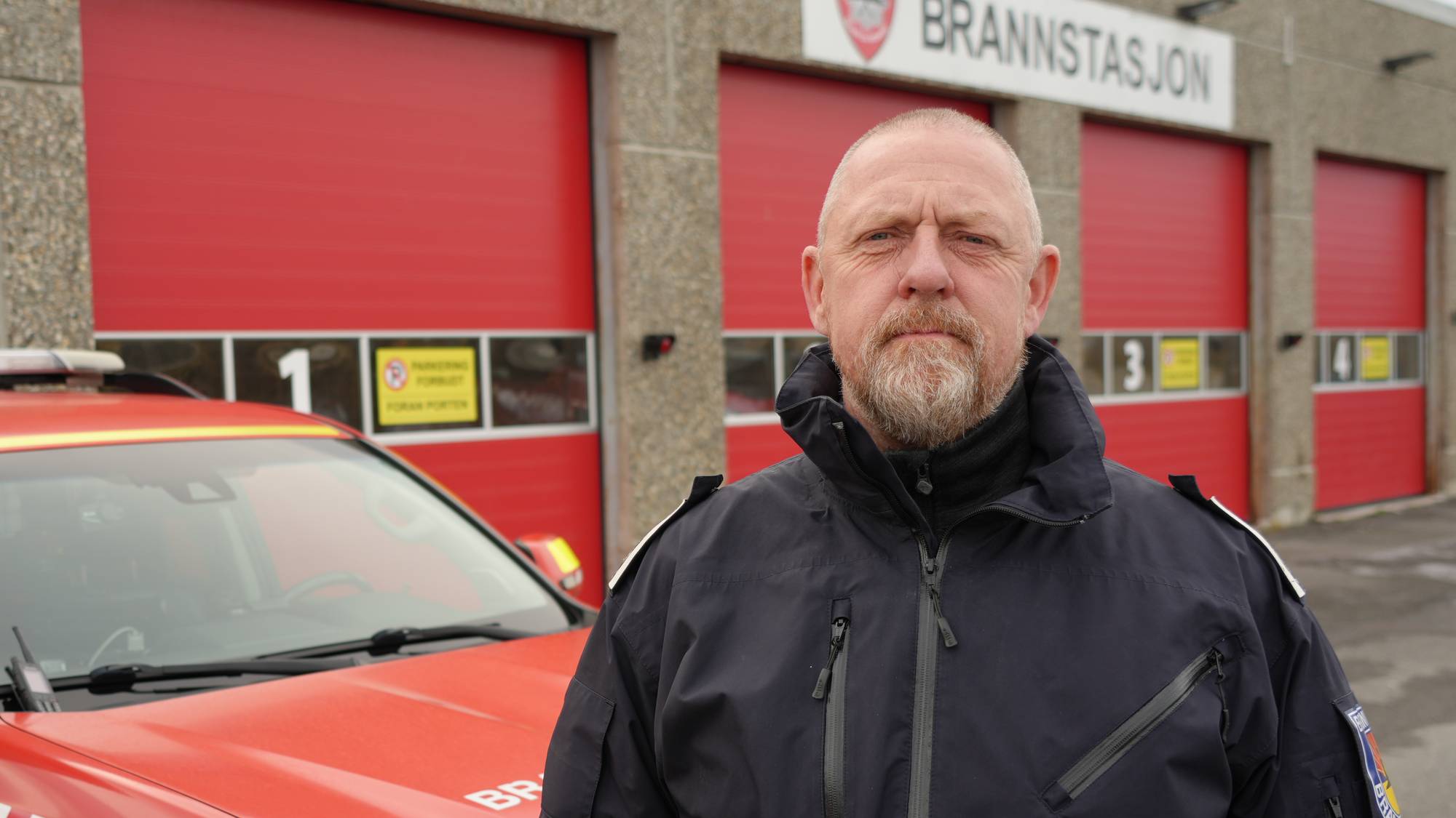 En innsatsleder for brannvesenet foran en brannstasjon med en rød utrykningsbil. 