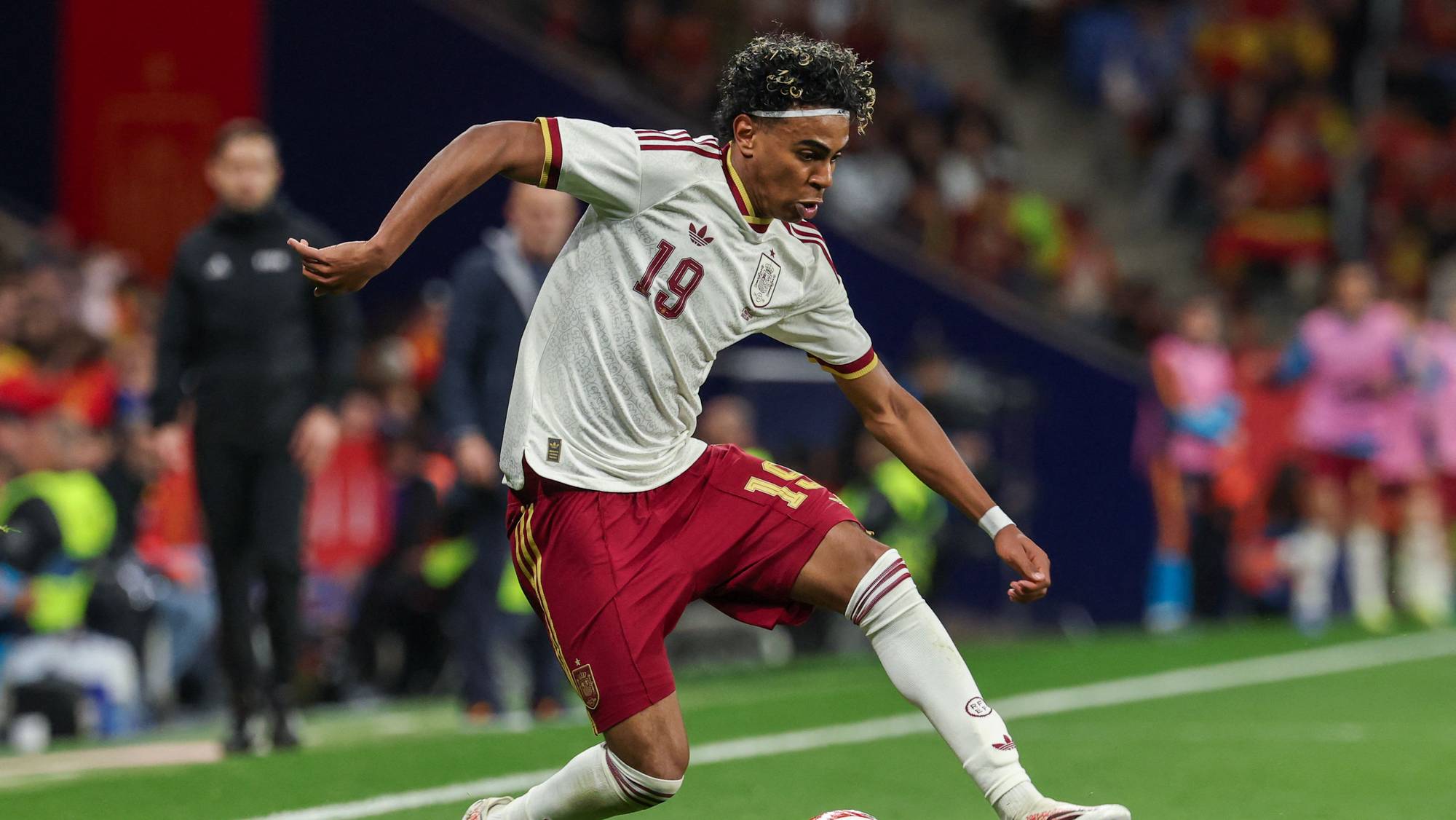 En fotballspiller med nummer 19 på drakten sparker til en fotball. Spilleren har på seg en hvit og burgunder drakt. I bakgrunnen sees publikum på en fullsatt tribune. (Bildebeskrivelsen er laget av en KI-tjeneste)
