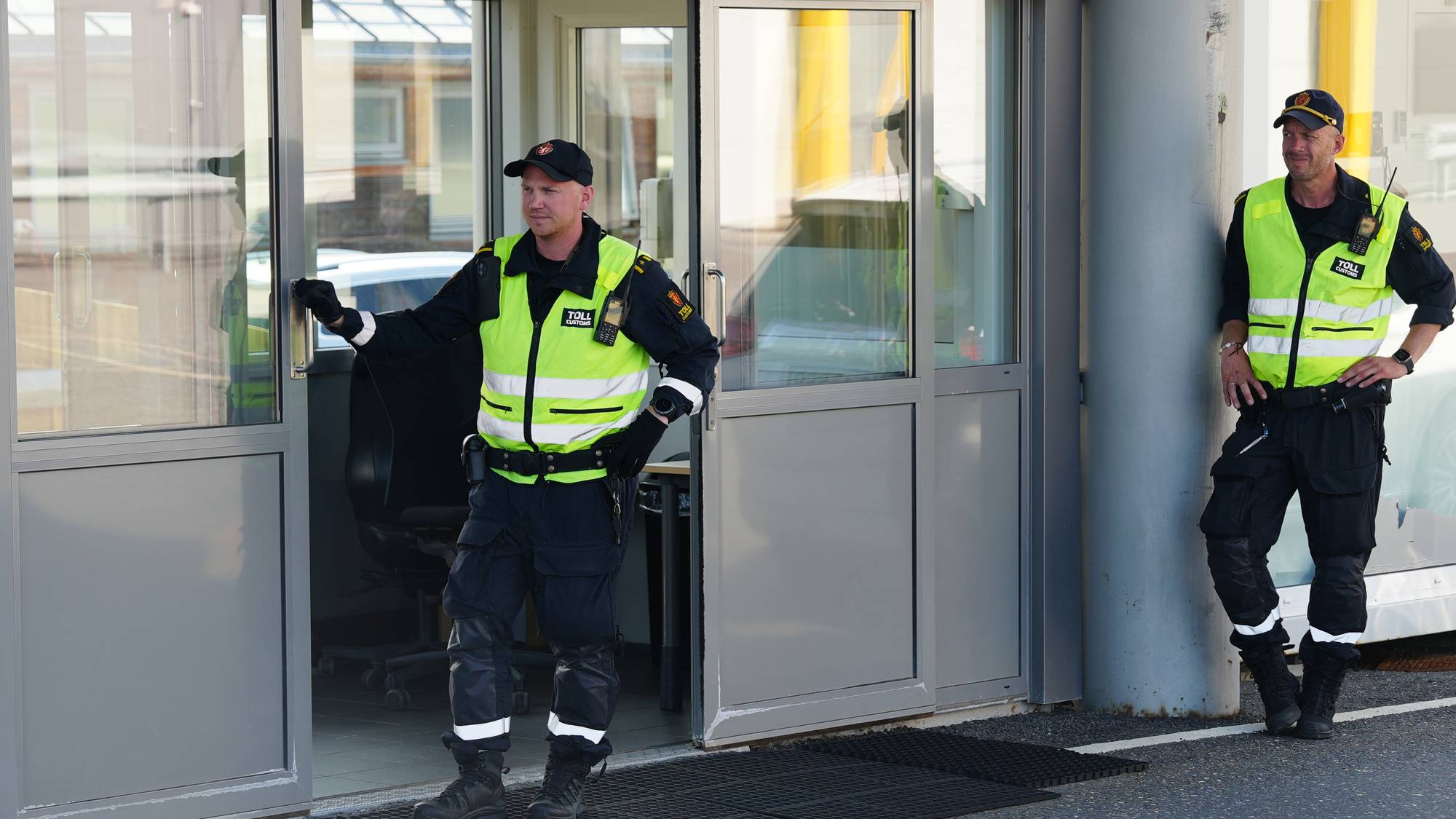 To tollbetjenter i uniform står ved en tollstasjon utendørs. 