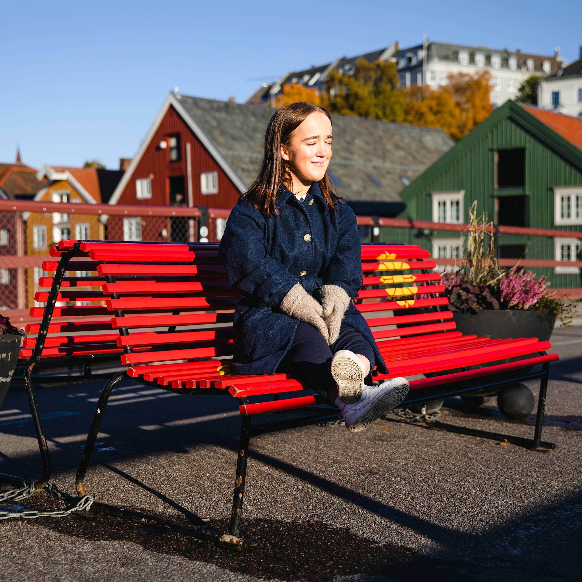 Oda sitter på en benk på broen ved Bakklandet og kniper øynene sammen mens hun smiler. Solen skinner. Bak henne ser vi den kjente utsikten til Trondheim ved Nideleven. 