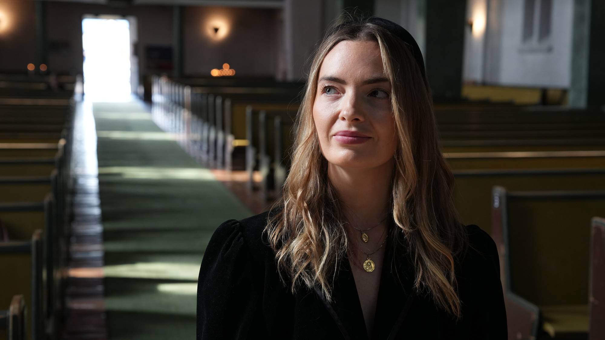Kvinne med mørke klær og smykker står i en kirke med rader av benker. (Bildebeskrivelsen er laget av en KI-tjeneste)
