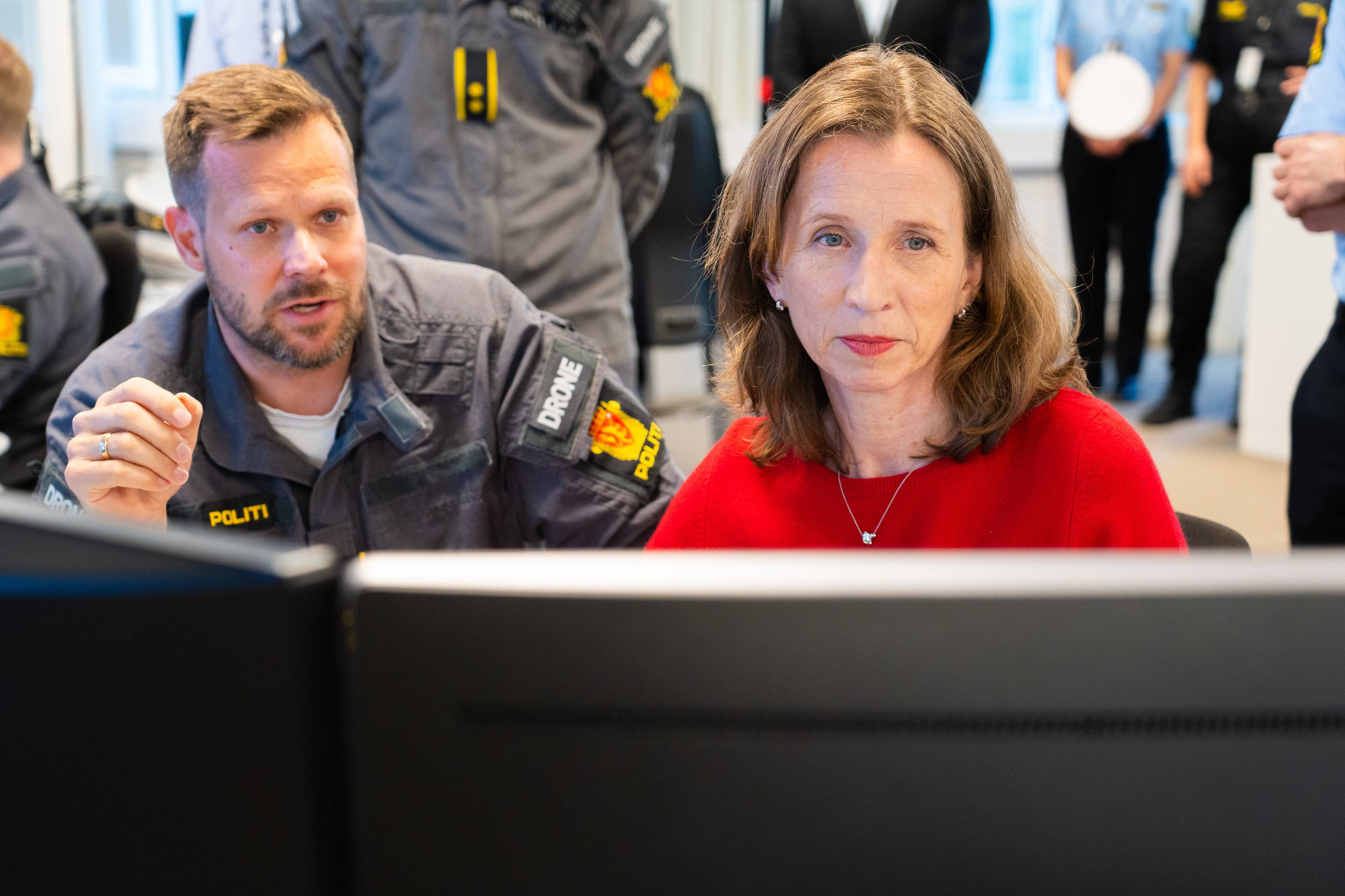 En mann i politiuniform snakker mens en kvinne i rød genser ser mot kamera. Bak dem står flere personer i uniform. De sitter foran dataskjermer. (Bildebeskrivelsen er laget av en KI-tjeneste)