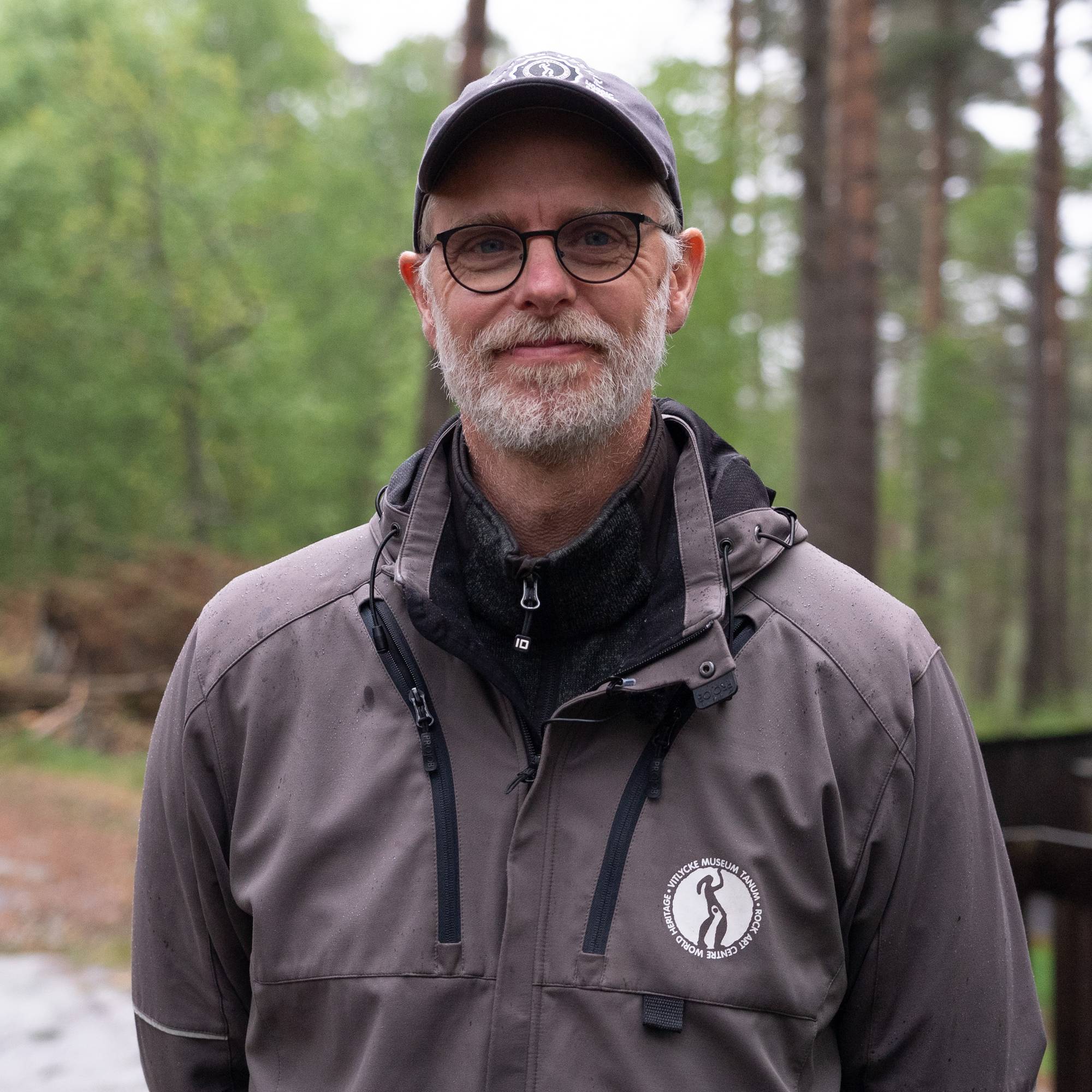 En mann med briller og caps står foran et skogholt. Han er kledd i en grå jakke med logo. Bak ham er det en skogssti og trær. Bildet er et fotografi. (Bildebeskrivelsen er laget av en KI-tjeneste)