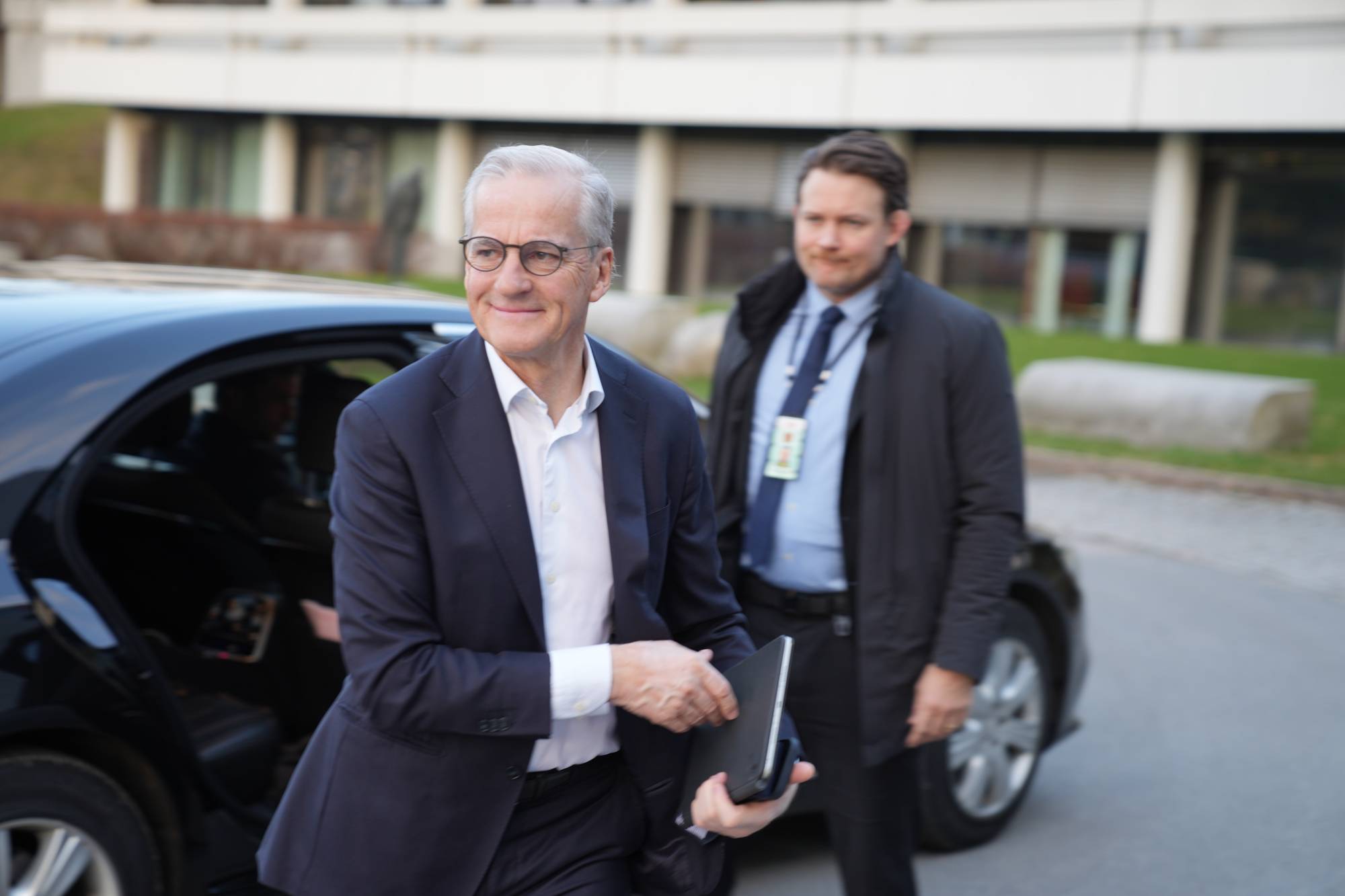 En mann i mørk dress går ut av en svart bil. Han smiler og holder en nettbrett. En annen mann i mørk jakke står bak ham. Bygningen bak dem er et moderne kontorbygg. (Bildebeskrivelsen er laget av en KI-tjeneste)