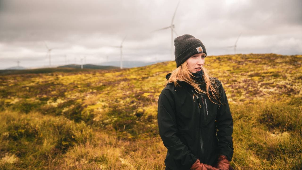 Et portrett av masterstudenten Aurora Ekker Jordahl, som står i myra i Roan vindkraftverk. Man skimter flere vindturbiner i bakgrunnen.