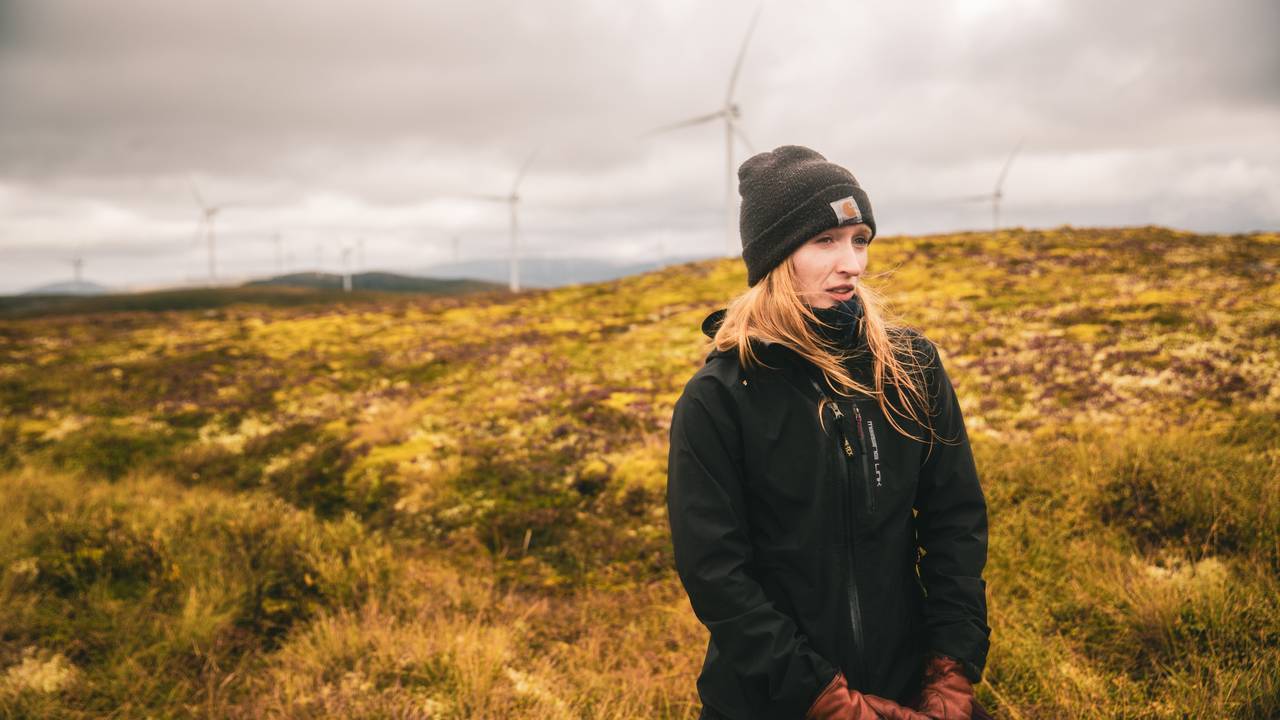Et portrett av masterstudenten Aurora Ekker Jordahl, som står i myra i Roan vindkraftverk. Man skimter flere vindturbiner i bakgrunnen.