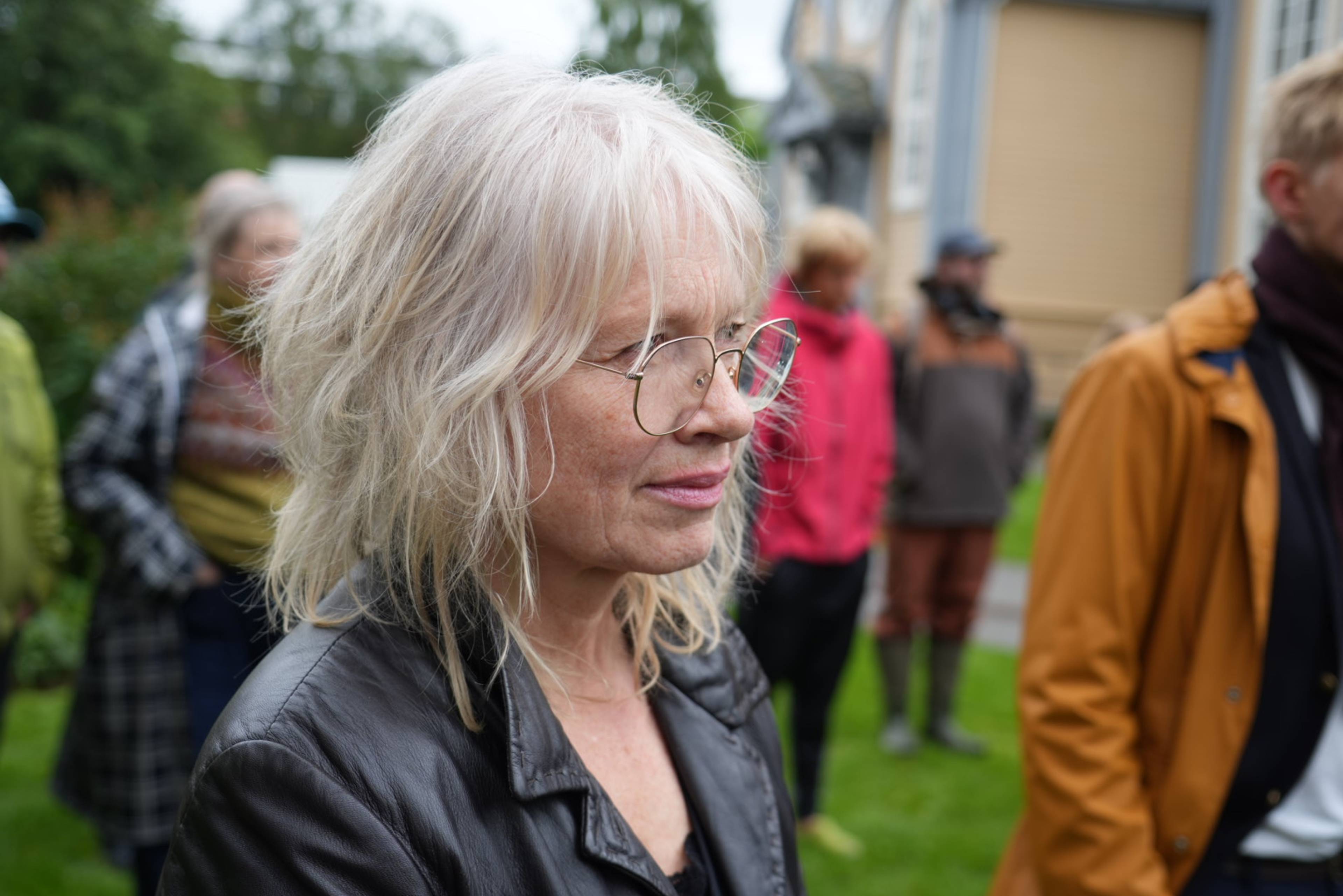 Kvinne med briller og lyst hår står utendørs med en folkemengde i bakgrunnen.