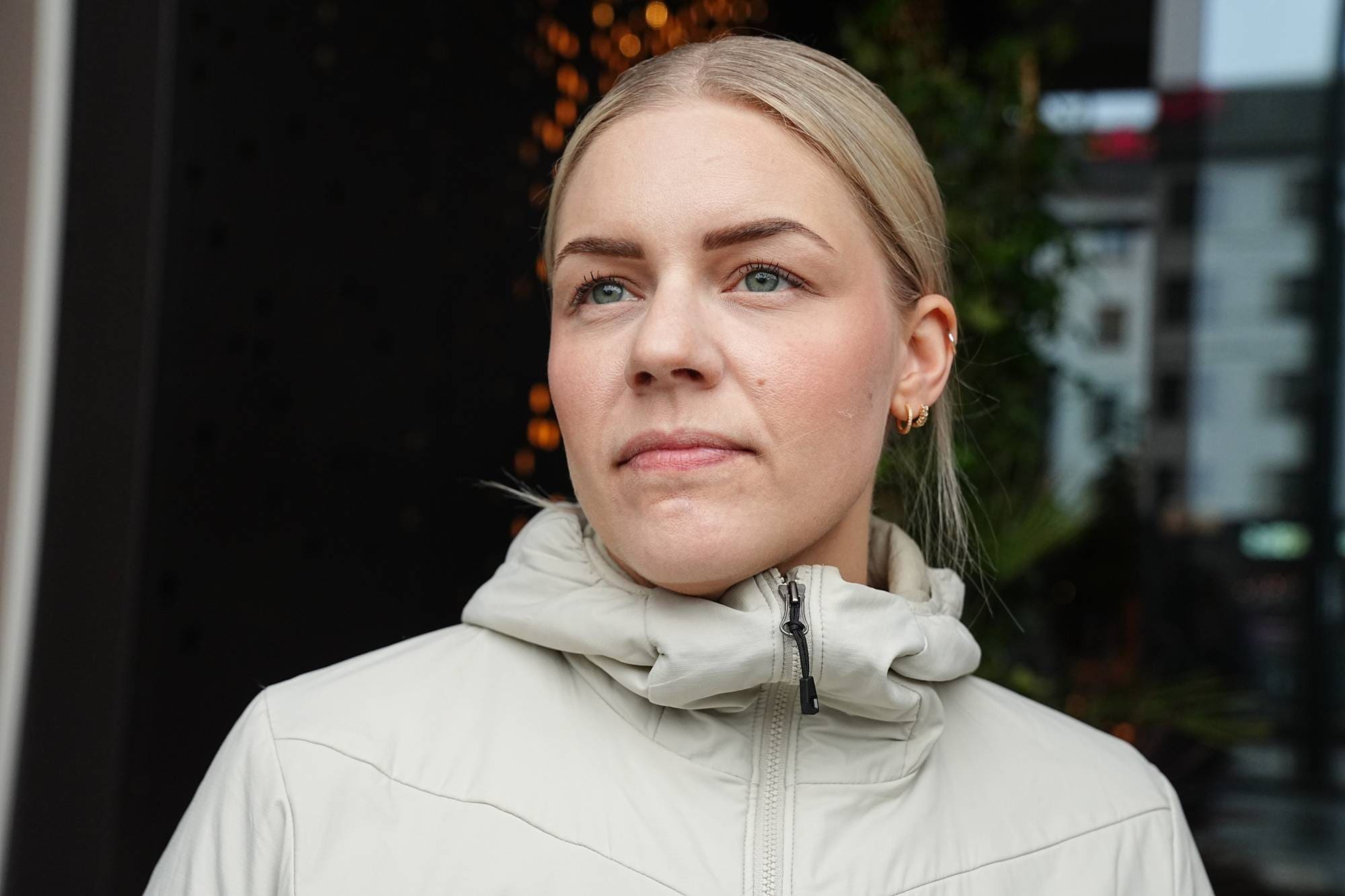 En kvinne med blondt hår ser til siden. Hun har på seg en lys jakke. Bak henne er det et mørkt parti med lysende dekorasjoner. (Bildebeskrivelsen er laget av en KI-tjeneste)