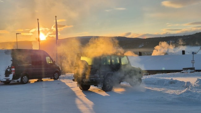 Et sort kjøretøy produserer damp i en snødekt parkeringsplass, mens solen går ned i bakgrunnen. I nærheten står en svart varebil delvis dekket av snø. I bakgrunnen kan man se bygninger med røykluer som slipper ut damp. To flaggstenger er synlige ved siden av varebilen. (Bildebeskrivelsen er laget av en KI-tjeneste)
