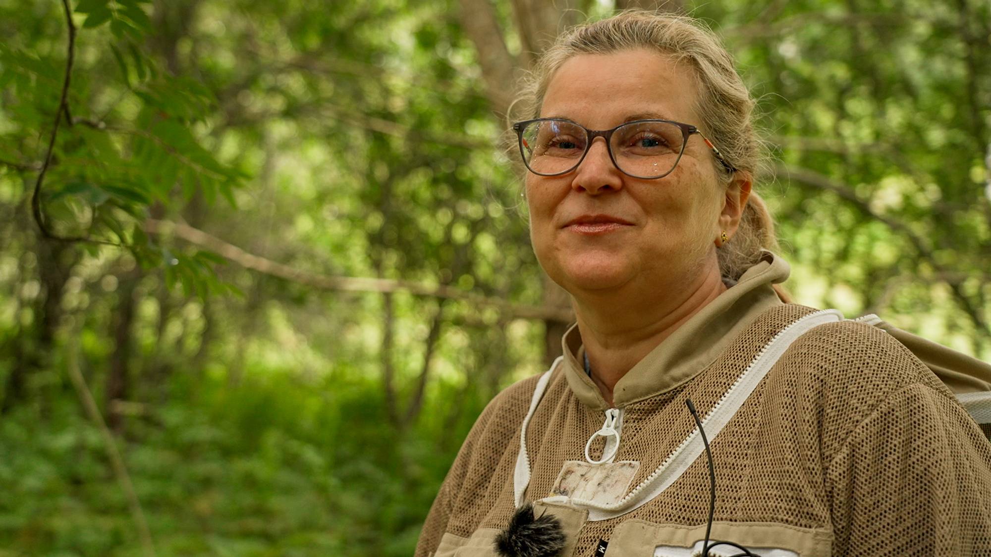 Kvinne med briller og birøkterdress står i en skog. (Bildebeskrivelsen er laget av en KI-tjeneste)