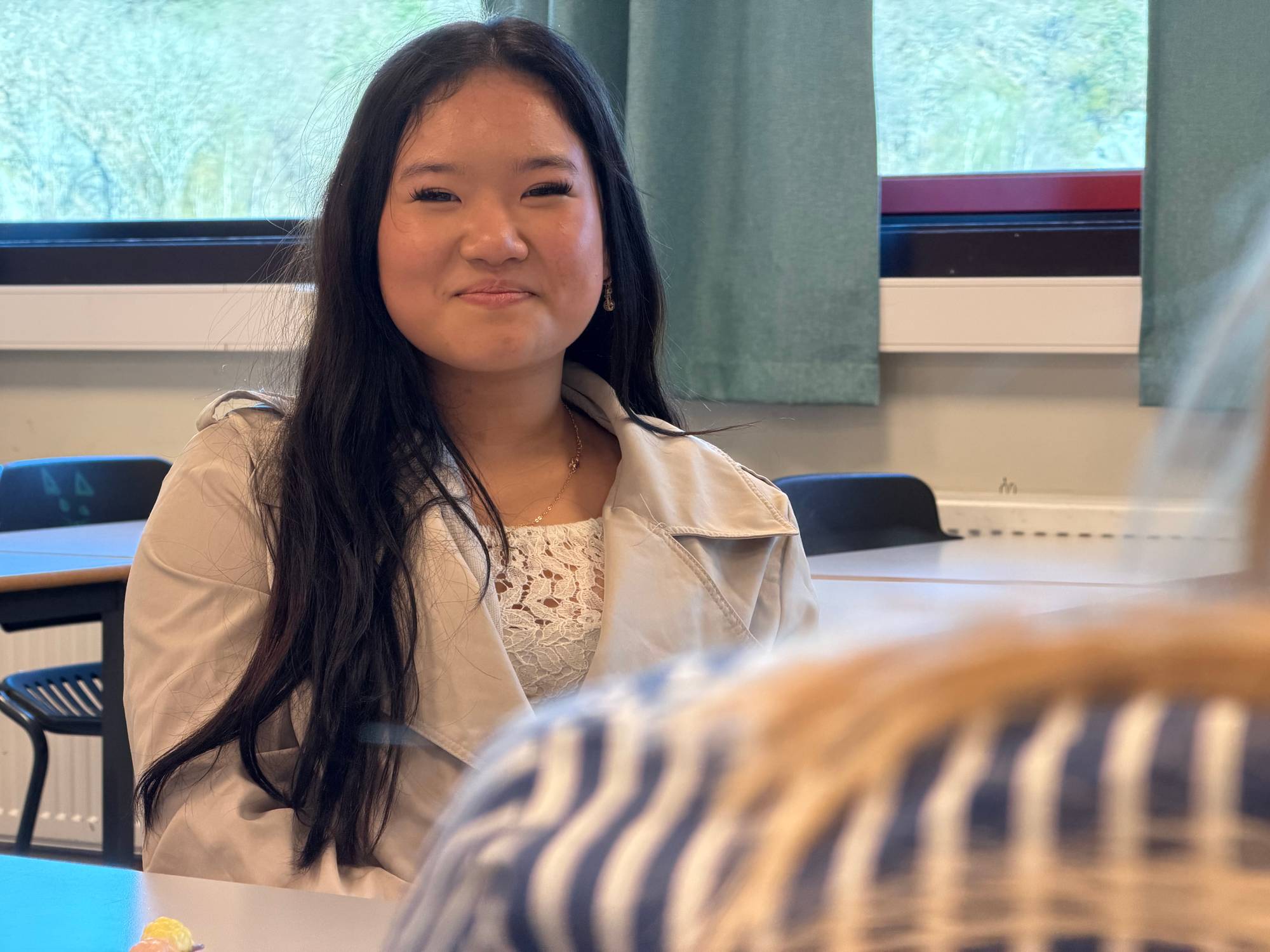 Ung kvinne sitter ved et bord. Hun har langt, mørkt hår og ser mot kamera. Bildet er tatt innendørs med et vindu i bakgrunnen. Det er en annen person foran. (Bildebeskrivelsen er laget av en KI-tjeneste)