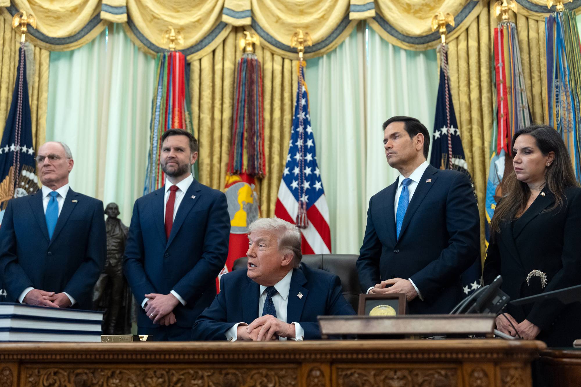 President Donald Trump sitter ved et skrivebord mens fem personer står bak ham. Trump har hendene på skrivebordet. Mennene til venstre står med hendene bak ryggen eller foran seg. Kvinnen til høyre står med hendene foran seg. I bakgrunnen er det et amerikansk flagg og flere andre flagg. (Bildebeskrivelsen er laget av en KI-tjeneste)
