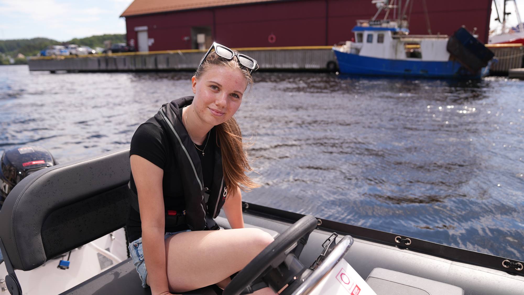 Ung kvinne med brunt langt hår og solbriller på sitter i gummibåt med rattet. Sjø og båt bak. 