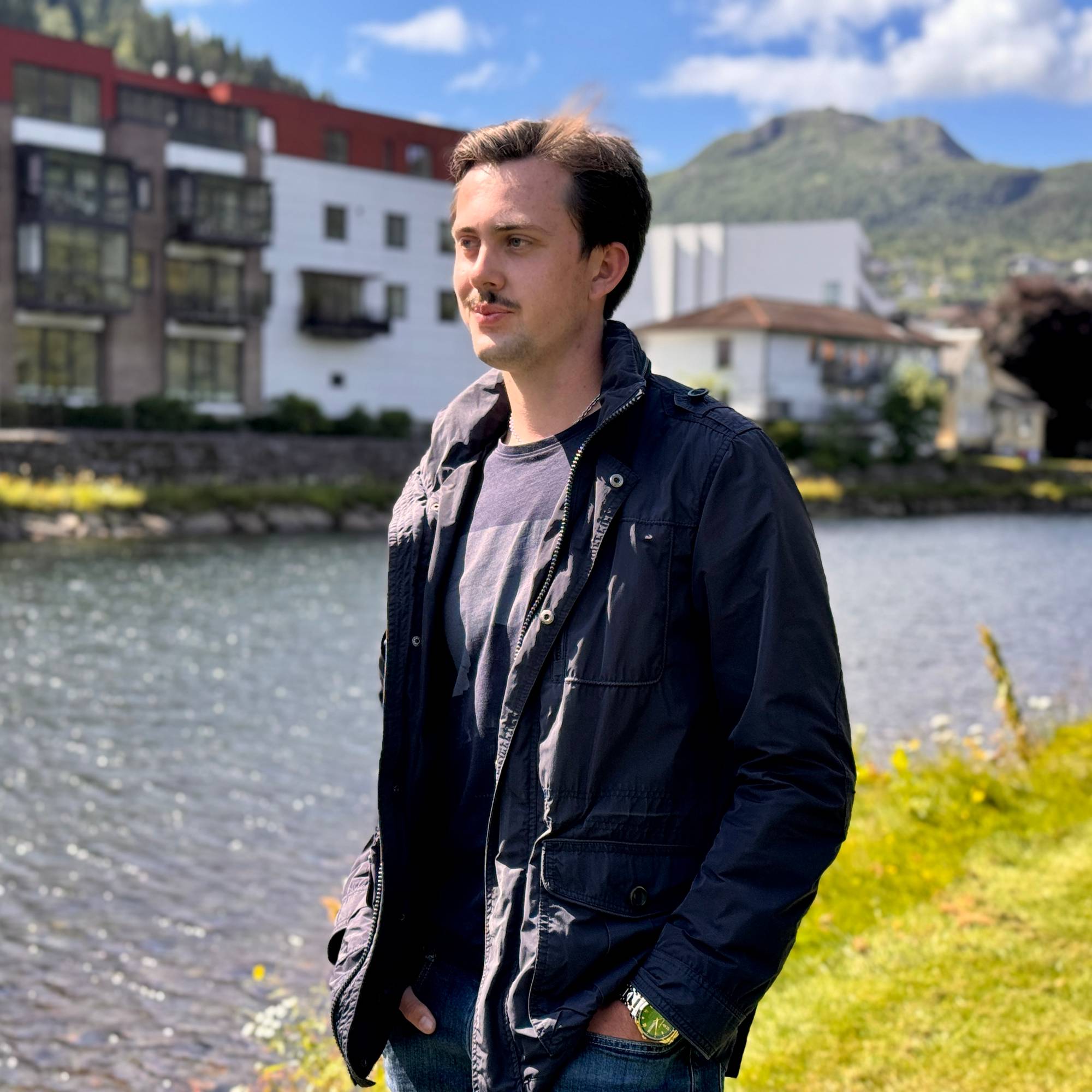 En ung mann står utendørs ved en elv med bebyggelse og fjell i bakgrunnen. (Bildebeskrivelsen er laget av en KI-tjeneste)