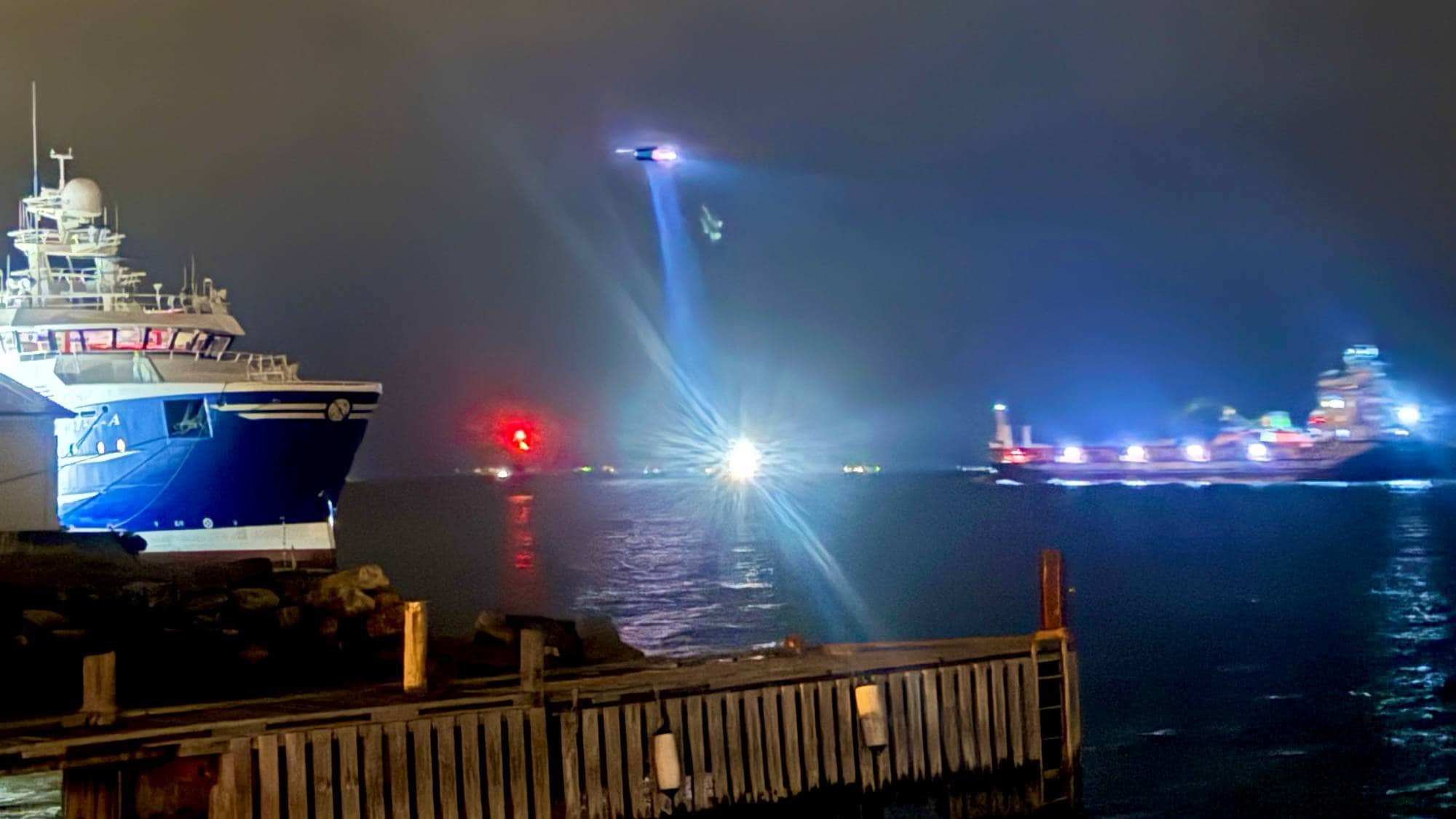 Et helikopter med lyskaster svever over sjøen mellom to skip om natten. En brygge er i forgrunnen. (Bildebeskrivelsen er laget av en KI-tjeneste)