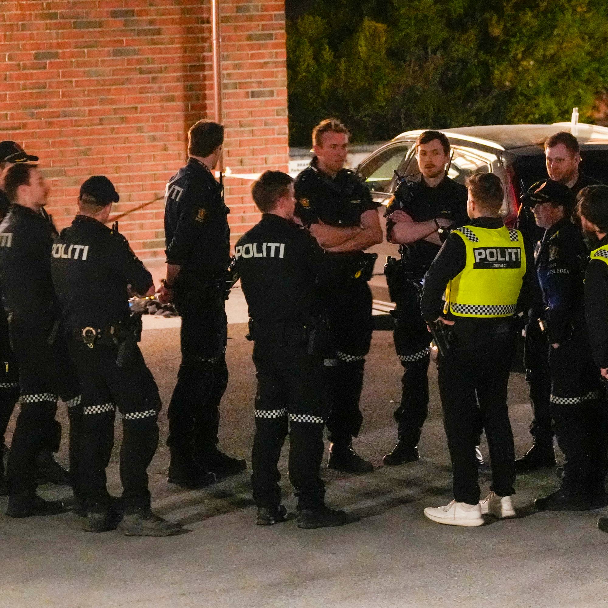 Politibetjenter er samlet utendørs foran en mursteinsbygning. Flere av dem er kledd i mørke uniformer med teksten "POLITI" på ryggen. Noen bærer gule refleksvester over uniformen. En bil står parkert i bakgrunnen. (Bildebeskrivelsen er laget av en KI-tjeneste)