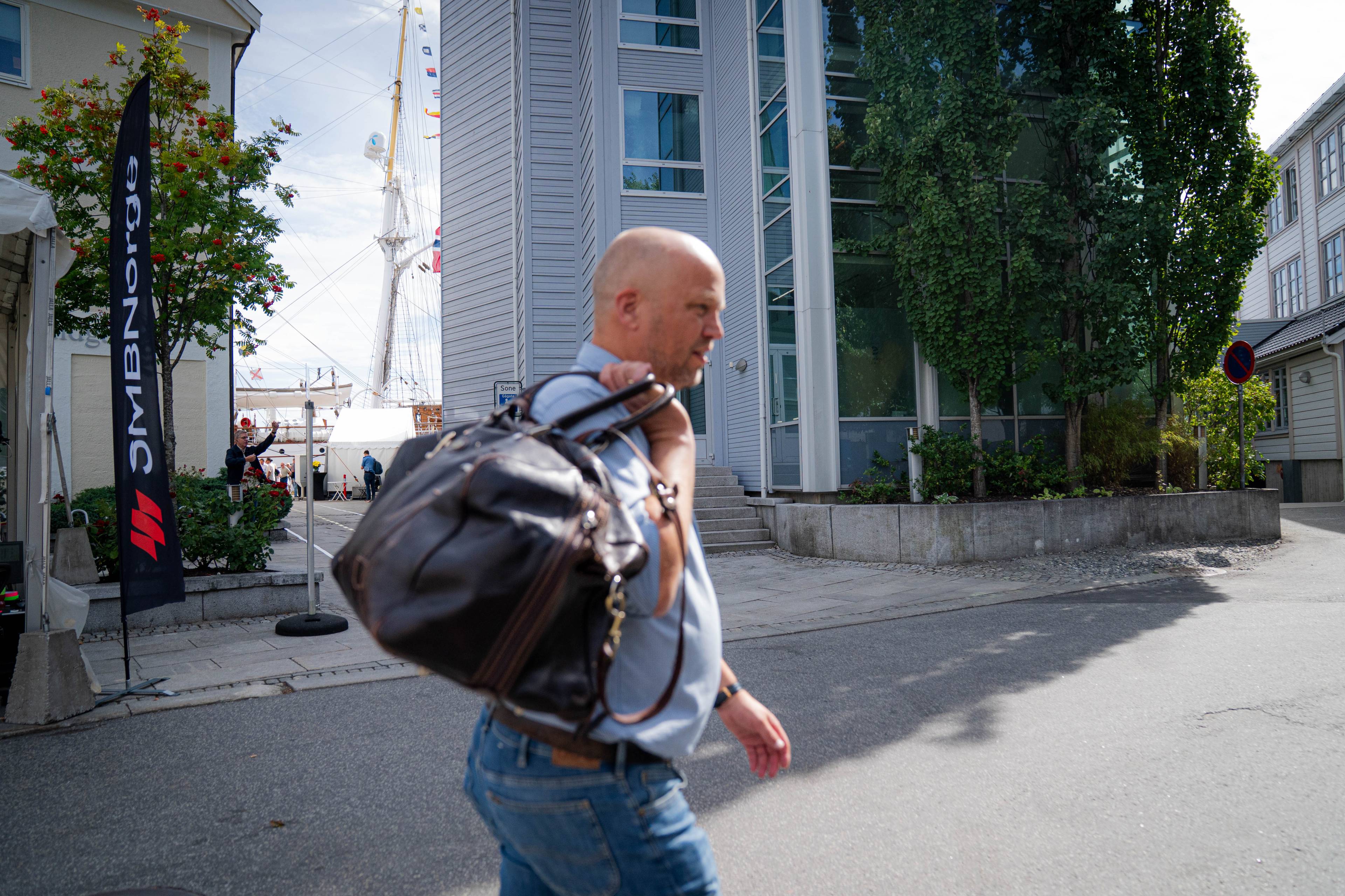 Skallet mann med skinnveske over skulderen går forbi moderne kontorbygg i et havneområde. (Bildebeskrivelsen er laget av en KI-tjeneste)