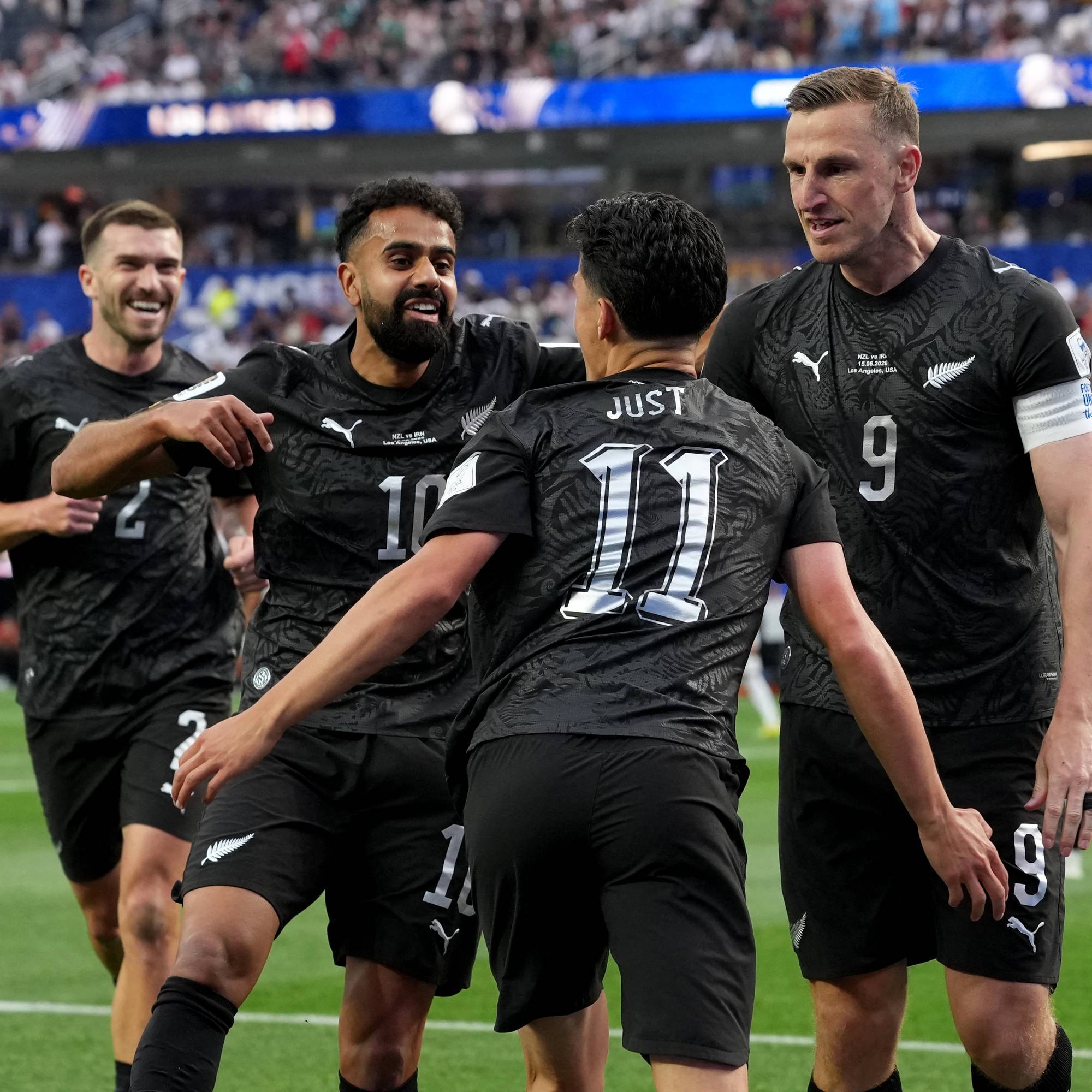 Fotballspillere i svarte drakter feirer på en gressbane. Flere spillere omfavner hverandre og smiler mens de ser mot siden. Tilskuere sitter på tribunen bak dem. (Bildebeskrivelsen er laget av en KI-tjeneste)