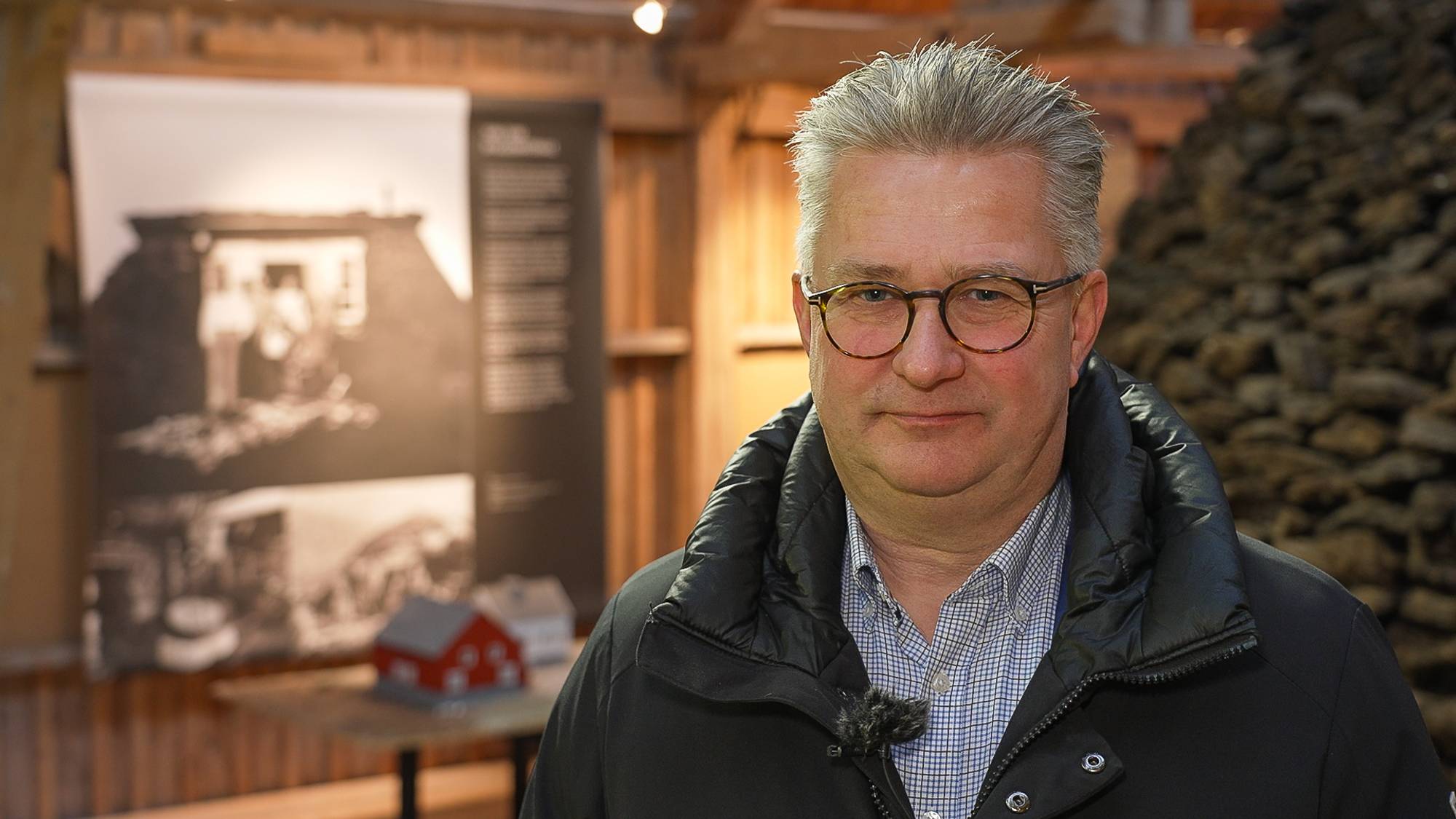 En mann står foran en utstilling i et rom med trevegger. I bakgrunnen finnes bilder av historiske bygninger og informasjonsplakater. Det ligger en liten modell av en rød bygning på et bord til venstre. Omgivelsene gir en følelse av kulturhistorisk kontekst.