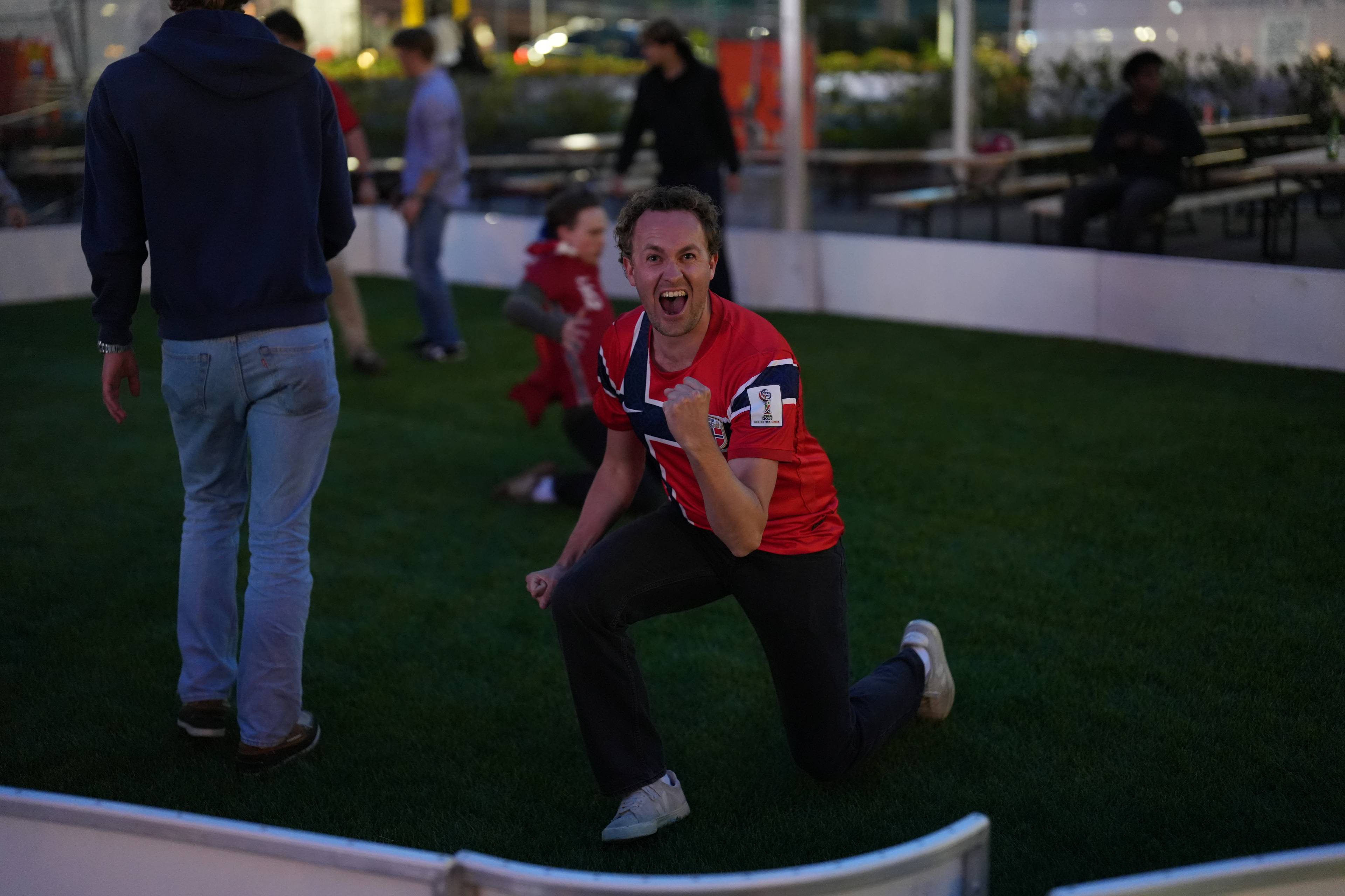 En mann i rød fotballdrakt med norsk flagg jubler. Han kneler på en kunstgressbane. Flere personer er i bakgrunnen.