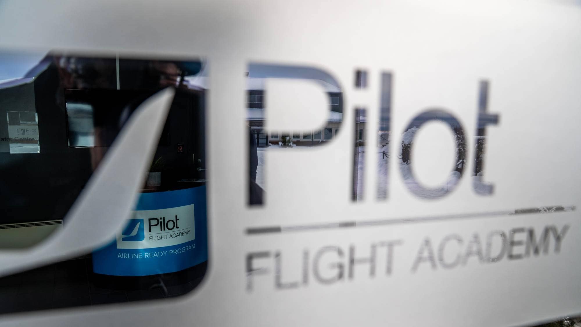 Bildet viser inngangen til Pilot Flight Academy, hvor logoen er synlig på en glassdør. Bak døren er en resepsjonsdisk med en blå bakgrunn med teksten "AIRLINE READY PROGRAM". Omgivelsene er preget av vinterlandskap utenfor, med snø som dekker bakken. Det er ikke synlige personer i bildet.