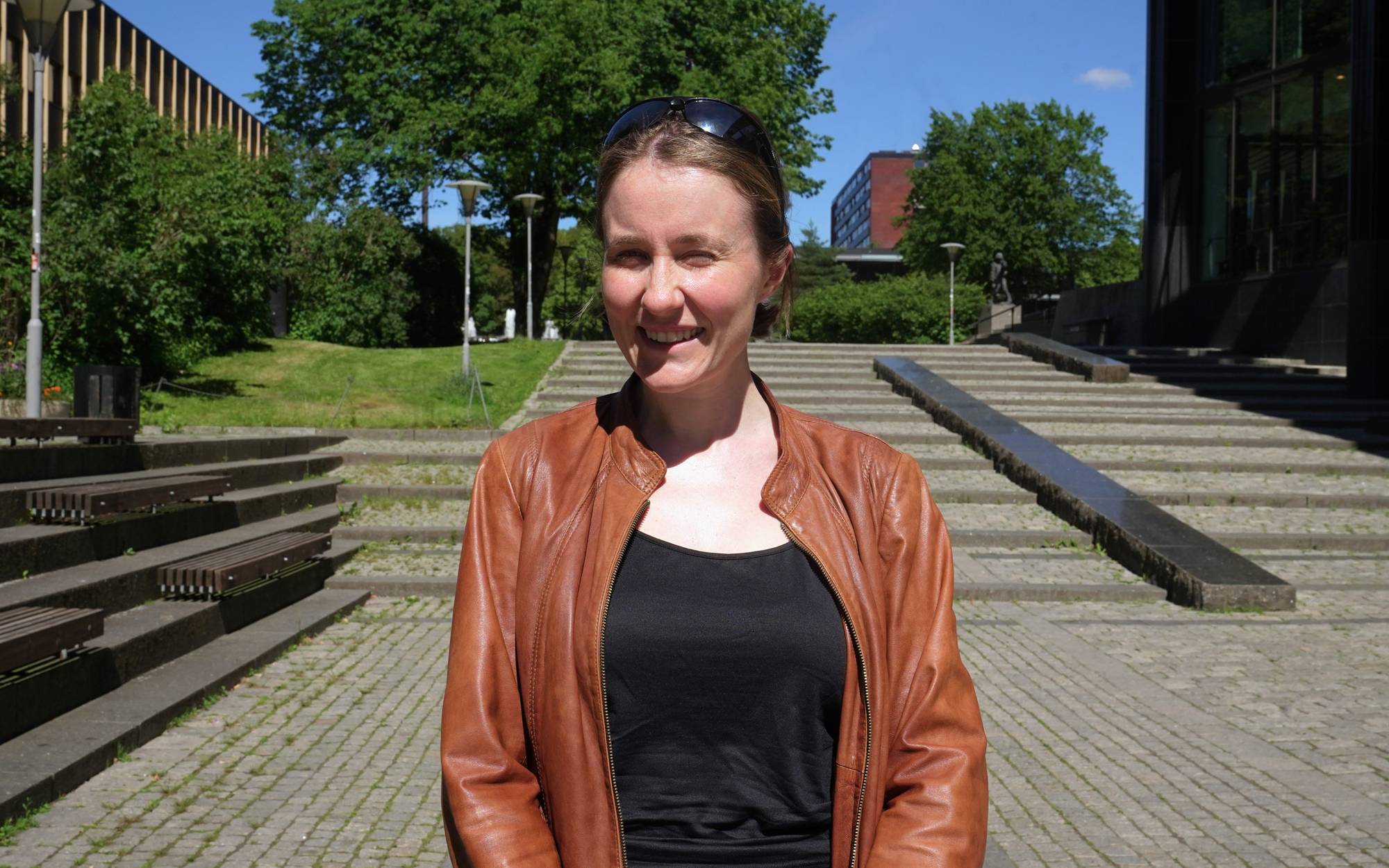 En kvinne med svart hopp og brun jakke smiler og myser litt i solen. Hun står utendørs ved et bibliotek på Blindern.