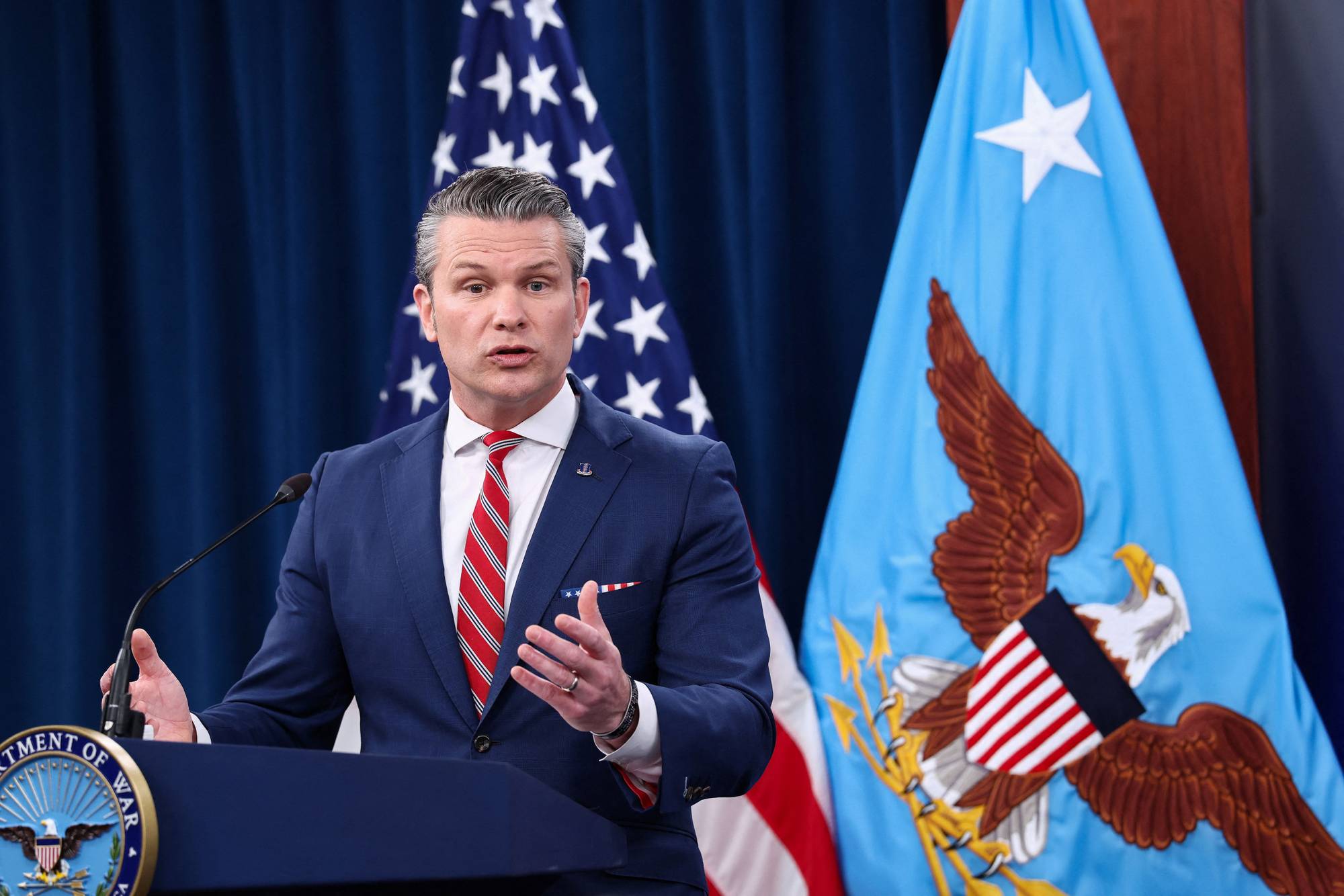 En mann i mørkeblå dress og rødt slips snakker bak et podium. Mannen står foran amerikanske flagg og et lyseblått flagg med en ørn. Bakgrunnen består av mørkeblå gardiner. (Bildebeskrivelsen er laget av en KI-tjeneste)