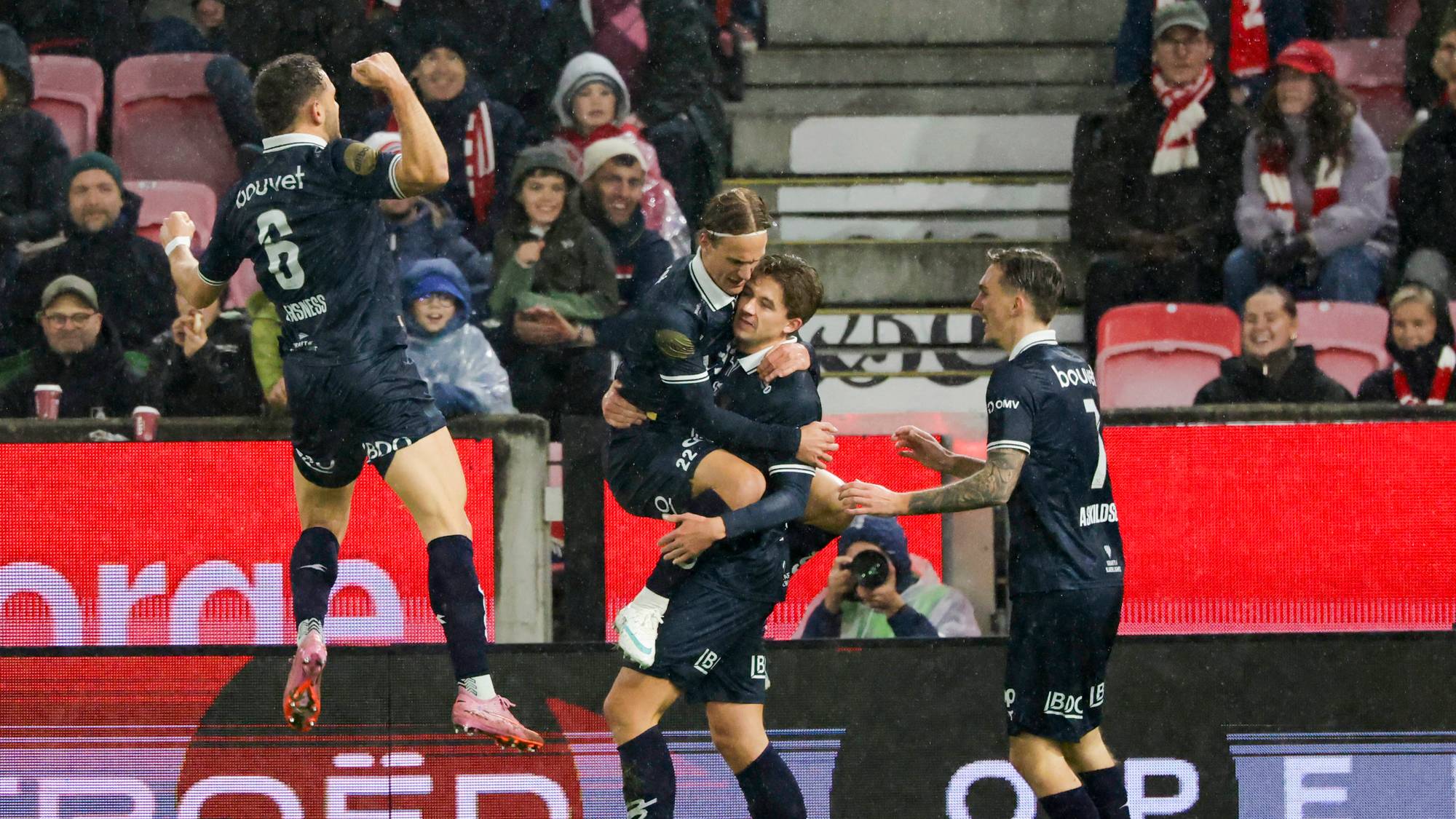 Fotballspillere i mørkeblå drakter jubler for mål på en fotballbane foran publikum. (Bildebeskrivelsen er laget av en KI-tjeneste)