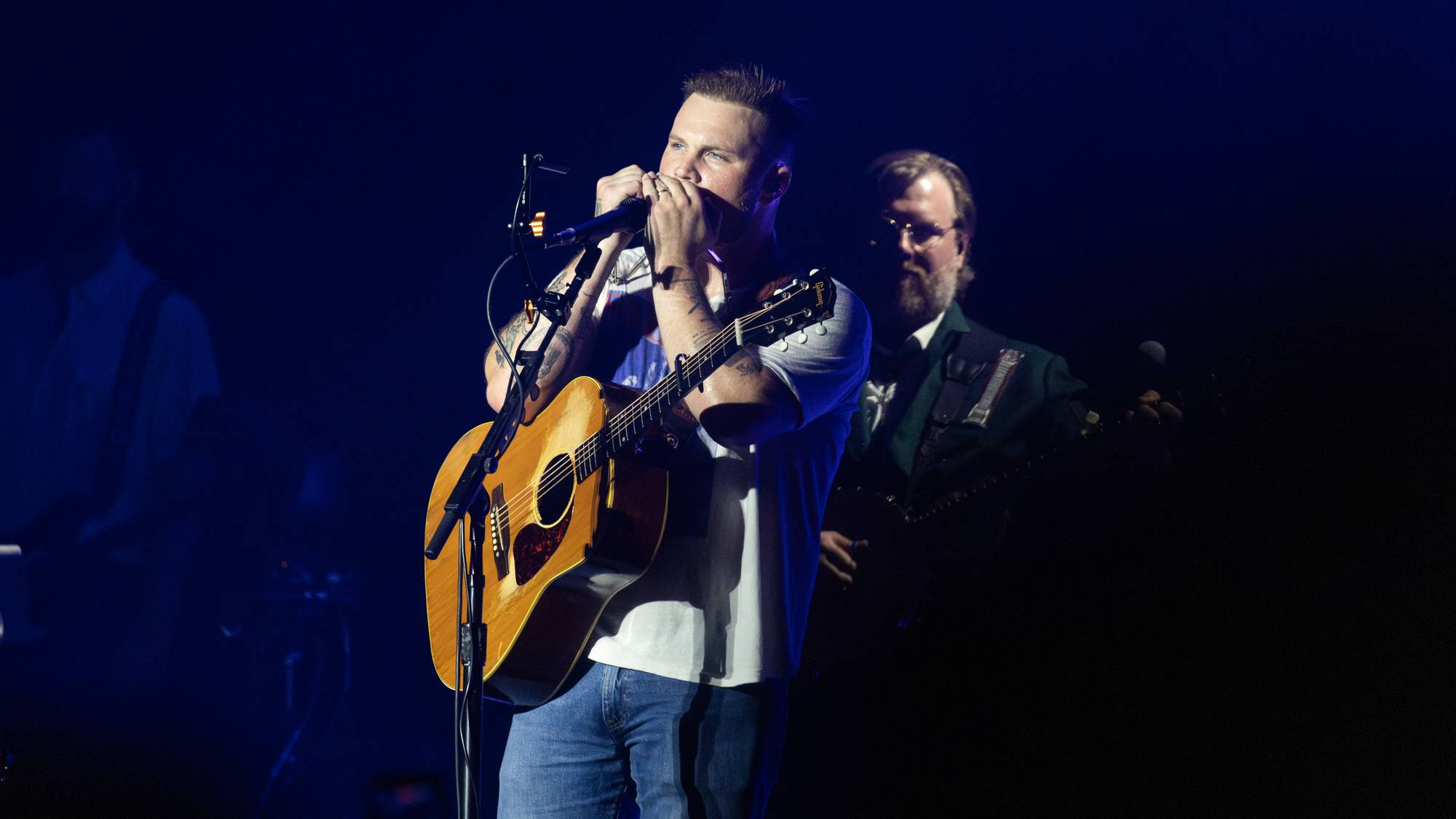 En mann spiller gitar og munnspill på en scene, mens en annen mann står bak. Mannen foran har på seg en hvit t-skjorte og jeans. Scenen er mørk, med blått lys som belyser artistene. (Bildebeskrivelsen er laget av en KI-tjeneste)