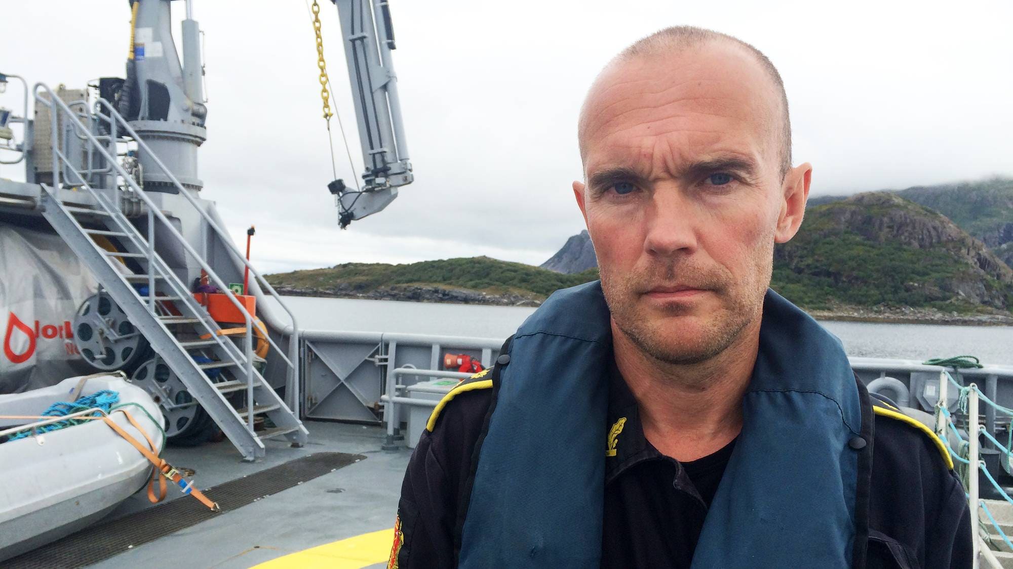 Politimann i uniform med redningsvest står på dekk på et fartøy med kystlandskap i bakgrunnen. (Bildebeskrivelsen er laget av en KI-tjeneste)