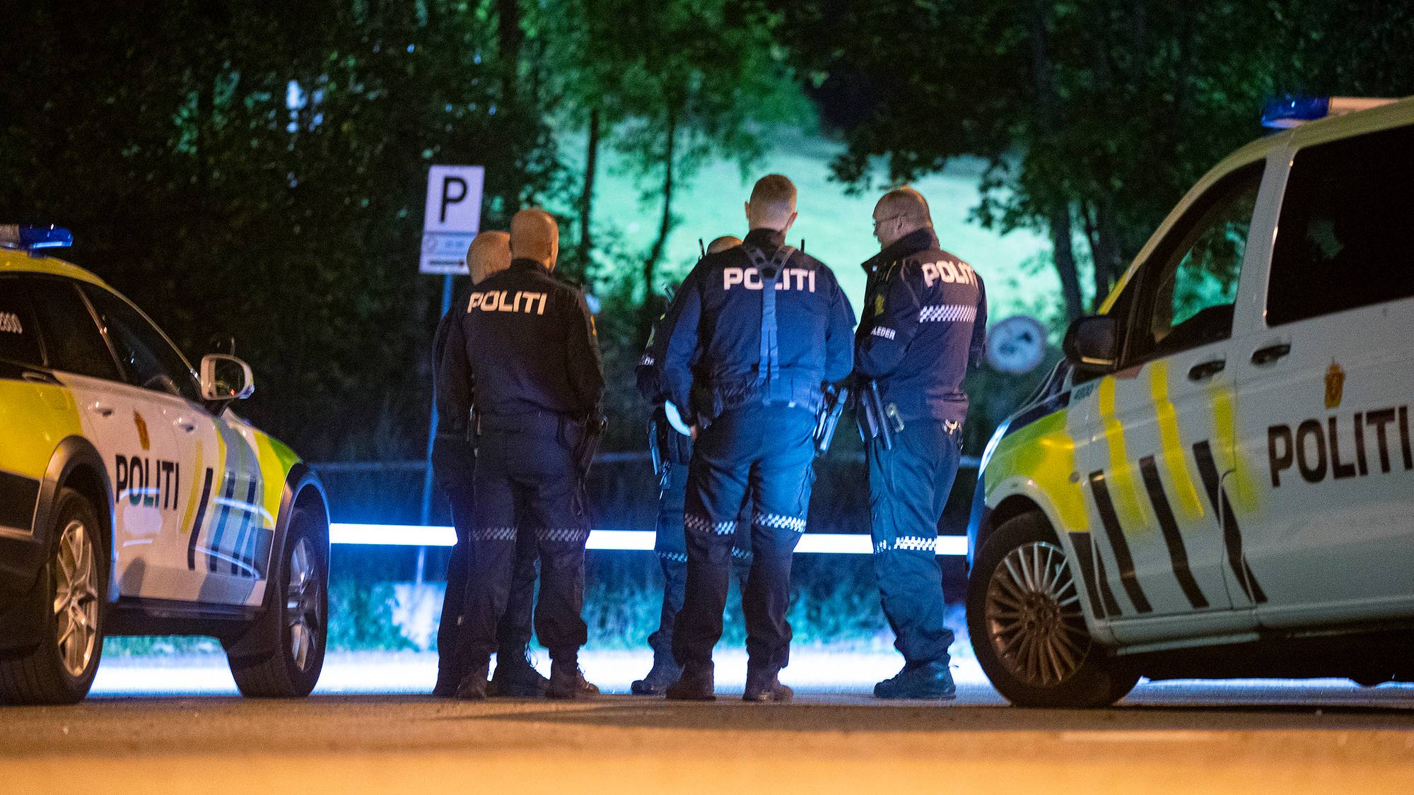 Flere MENNESKER MED UNIFORM DET STÅR "POLITI" på ryggen på står og diskuterer på et mørkt området. På hver side av dem står det en politibil som lyser opp veien foran dem. 