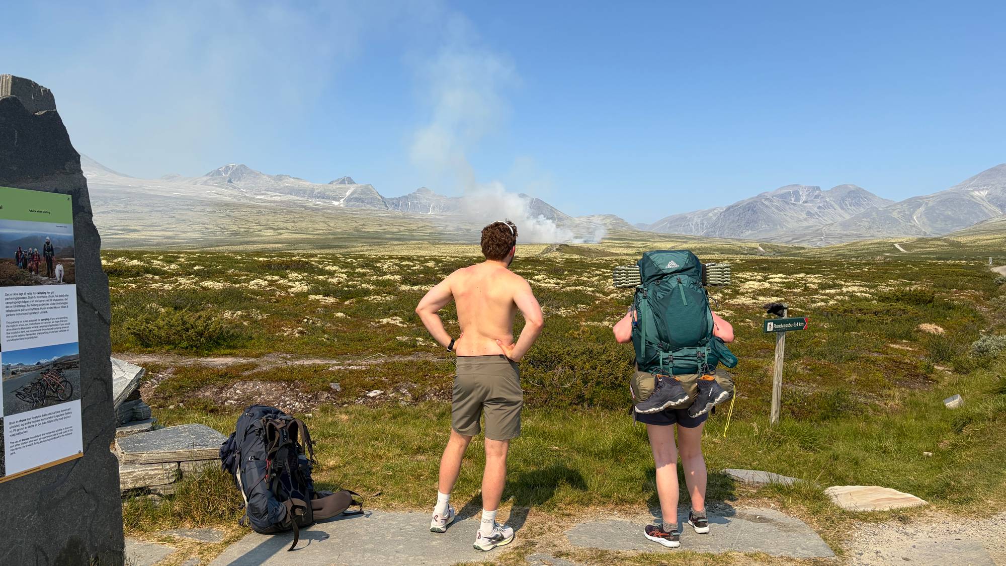 To personer med ryggsekker ser mot røykutvikling i et fjellandskap i Rondane. (Bildebeskrivelsen er laget av en KI-tjeneste)