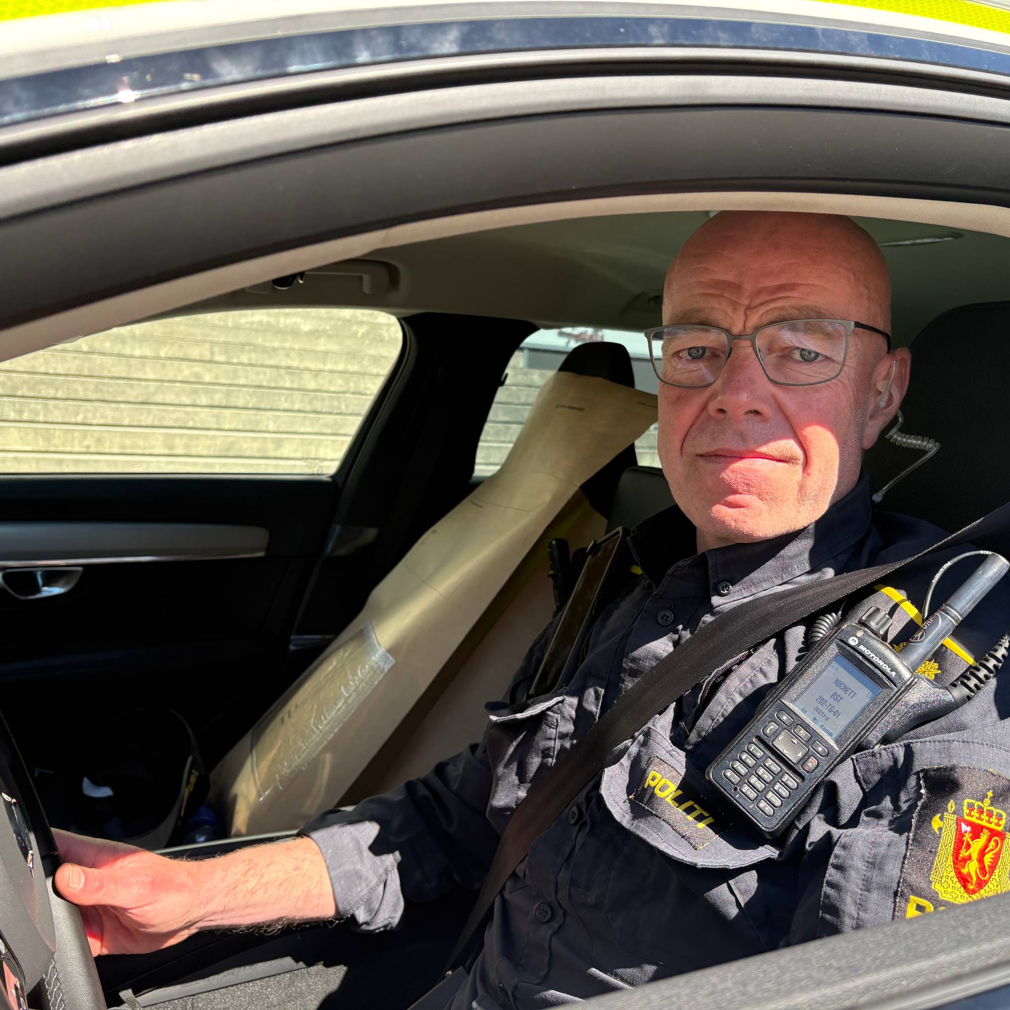 En mann i politiuniform sitter og ser ut inne i en politibil med nedrullet vindu. 