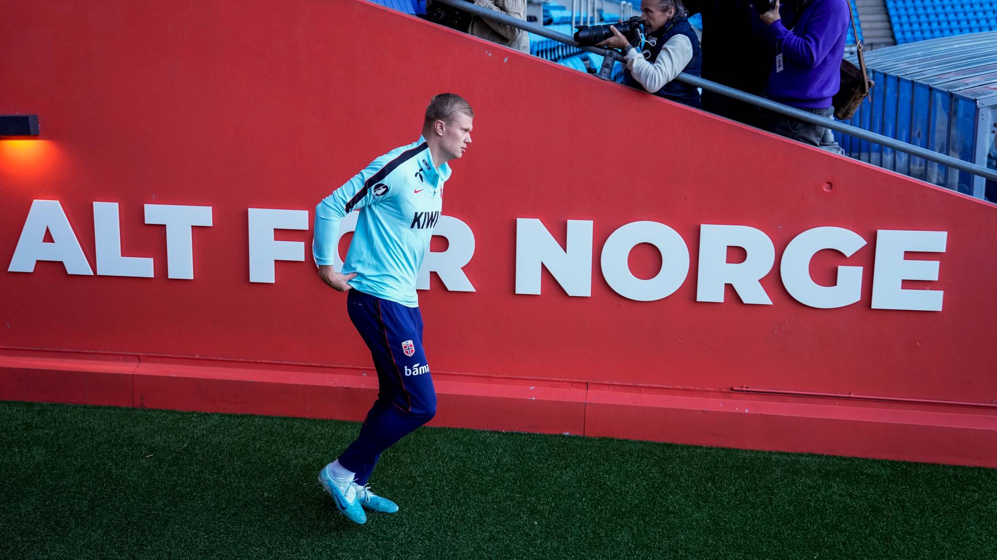 Erling Braut Haaland går forbi en rød vegg med teksten Alt for Norge på en fotballarena. (Bildebeskrivelsen er laget av en KI-tjeneste)
