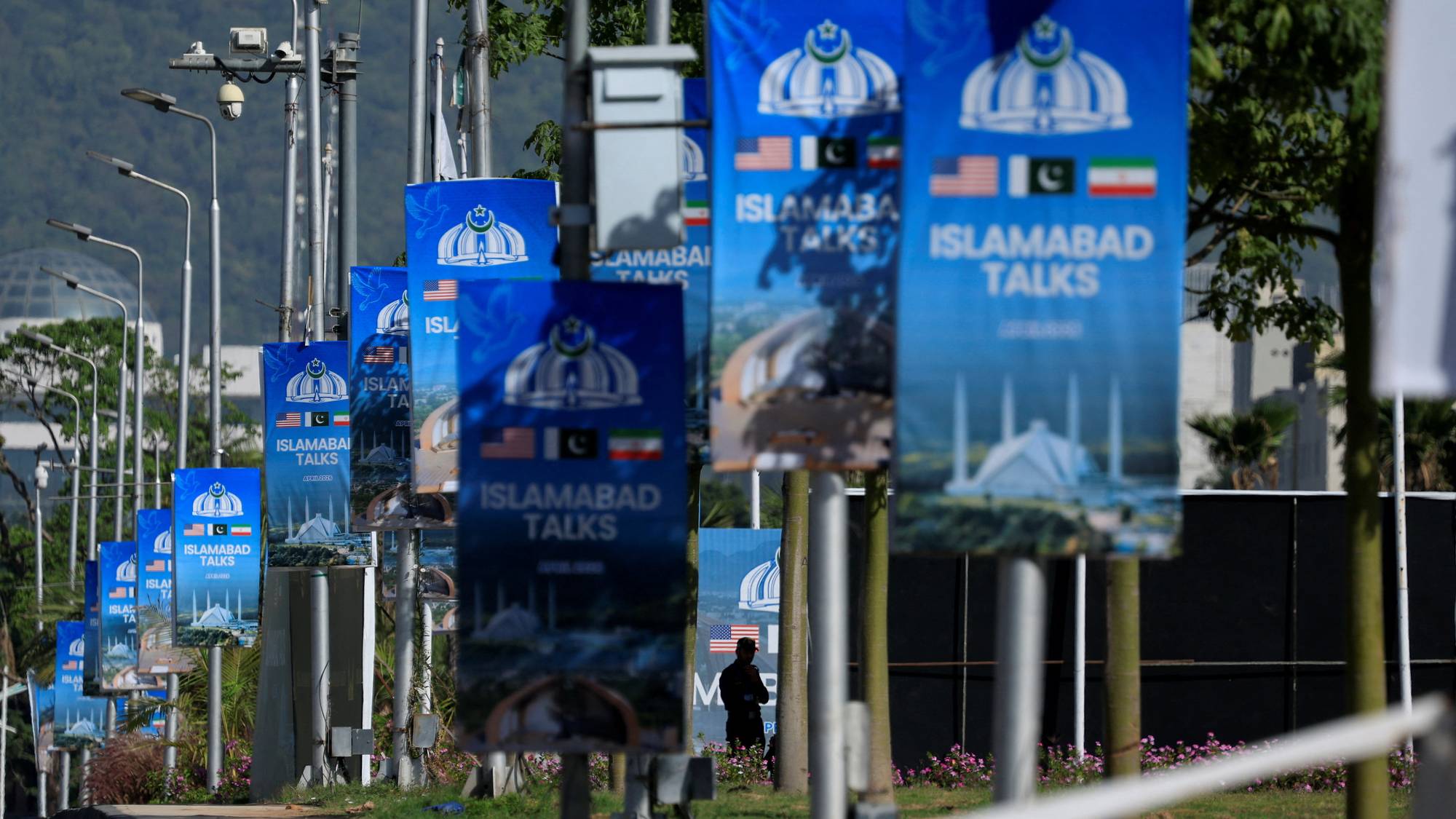 En rekke blå bannere med teksten "Islamabad Talks" henger langs en vei. Bannerne viser nasjonalflagg, et bilde av en moské og en halvmåne med en stjerne. I bakgrunnen sees trær og en ås. En mann står i silhuett mellom bannerne. (Bildebeskrivelsen er laget av en KI-tjeneste)
