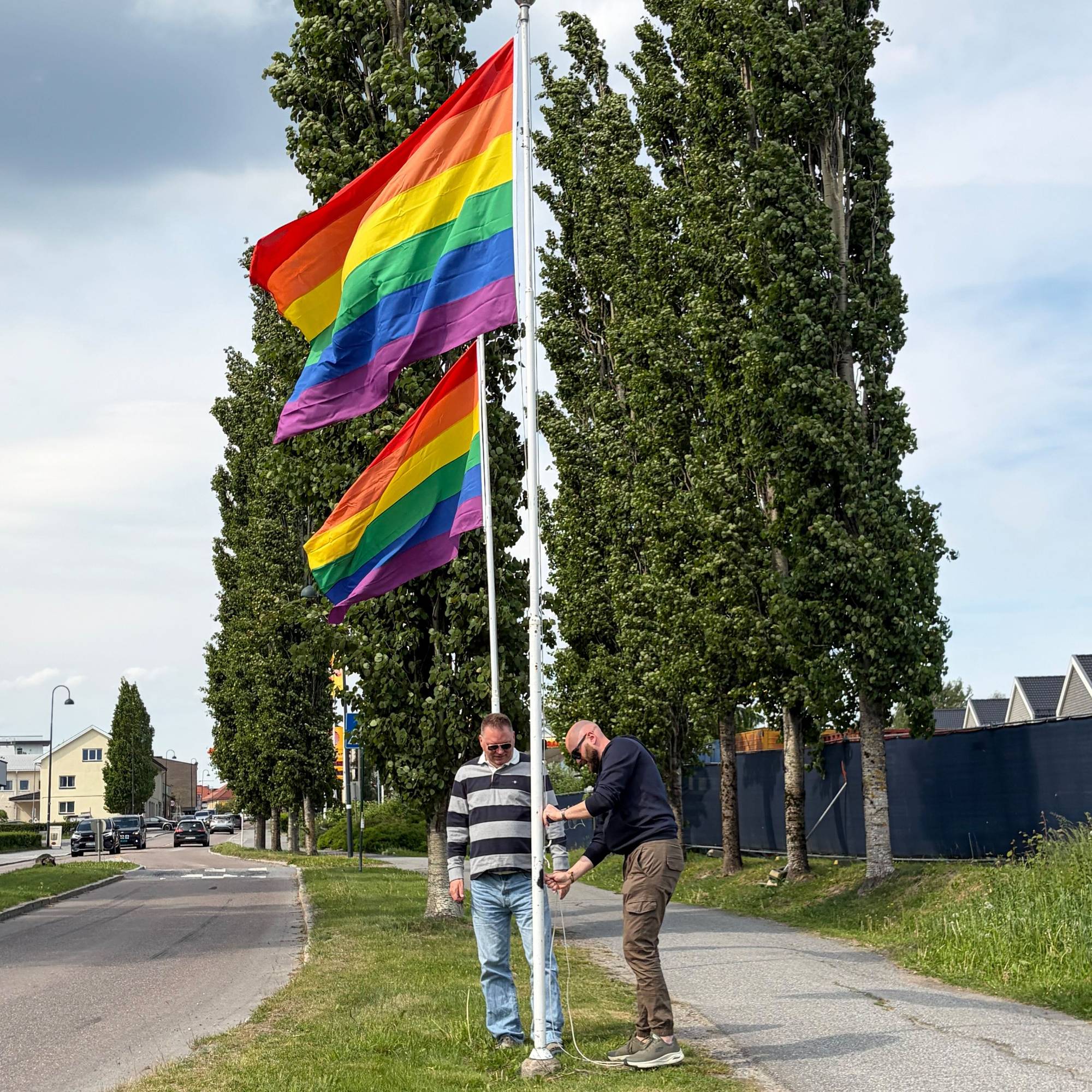 Prideflagg på rekke og rad i Rakkestad. To menn står ved en av flaggstengende hvor et prideflagg vaier i vinden. 
Flagget har striper i regnbuens farger: Rød, oransje, gul, grønn, blå og fiolett. 