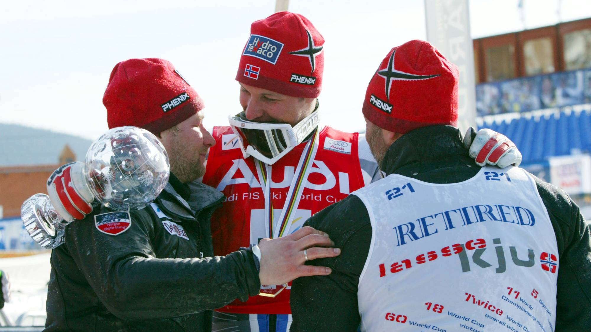 En mann med alpinbriller og medaljer gratuleres av to menn med røde luer. Den ene mannen holder et globuslignende trofé. Mannen i midten har en rød jakke med logoer. (Bildebeskrivelsen er laget av en KI-tjeneste)