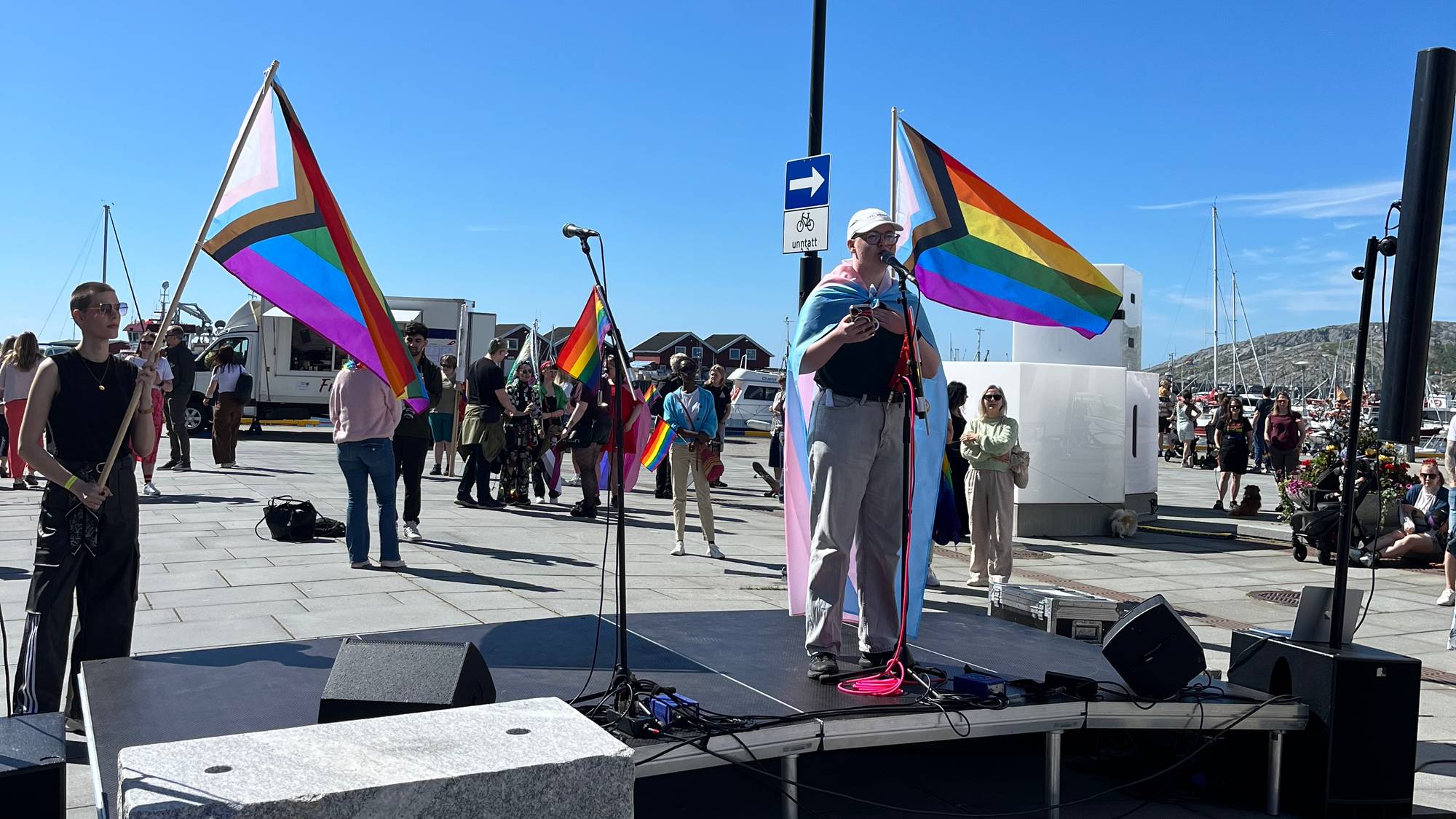 Leder for FRI Nordland, Luca Nielsen Jenssen, står på en liten sene og holder tale under pridemarkeringen i Bodø. På hver ide av ham står det en person med et prideflagg.